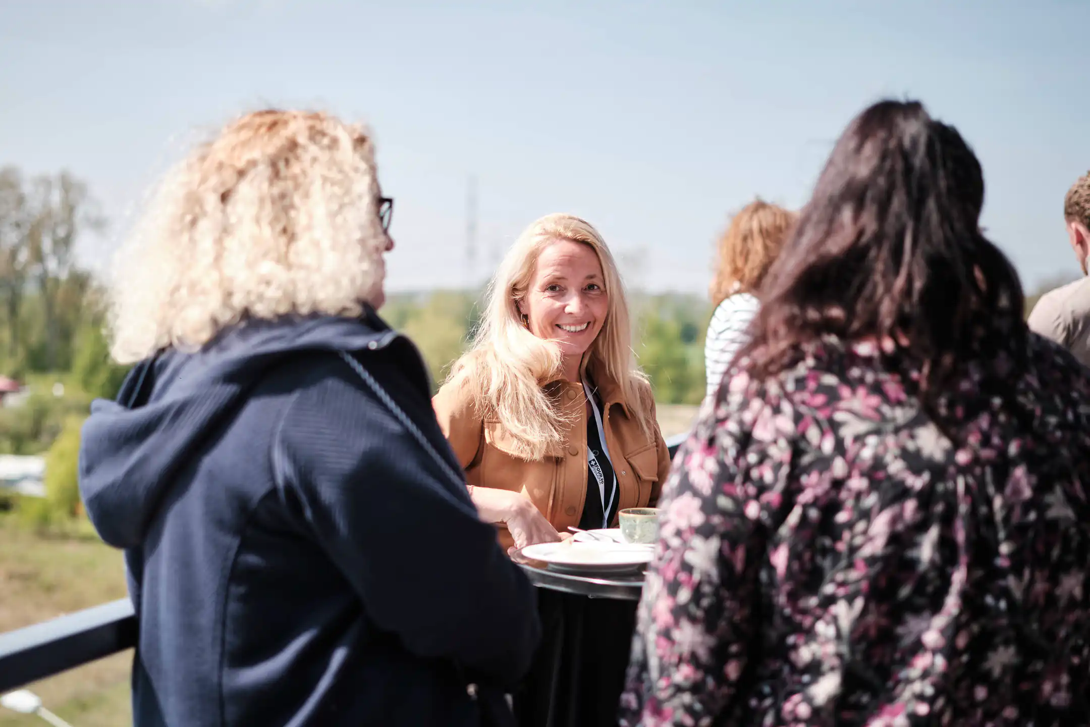 Drei Frauen unterhalten sich draußen, eine blonde Frau lächelt in die Kamera bei sonnigem Wetter.