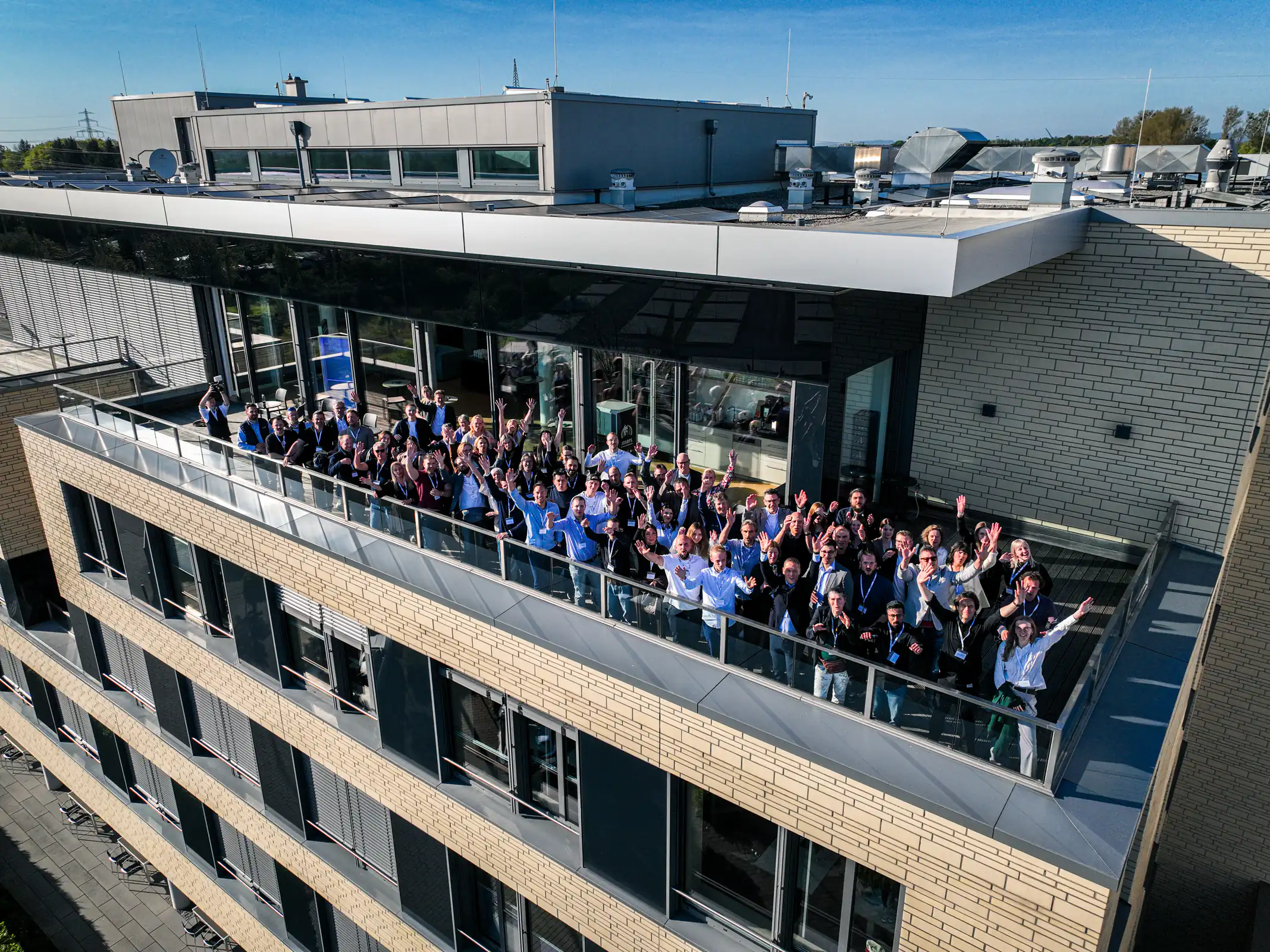 Große Gruppe Menschen winkt auf Balkon eines modernen Bürogebäudes bei sonnigem Wetter.