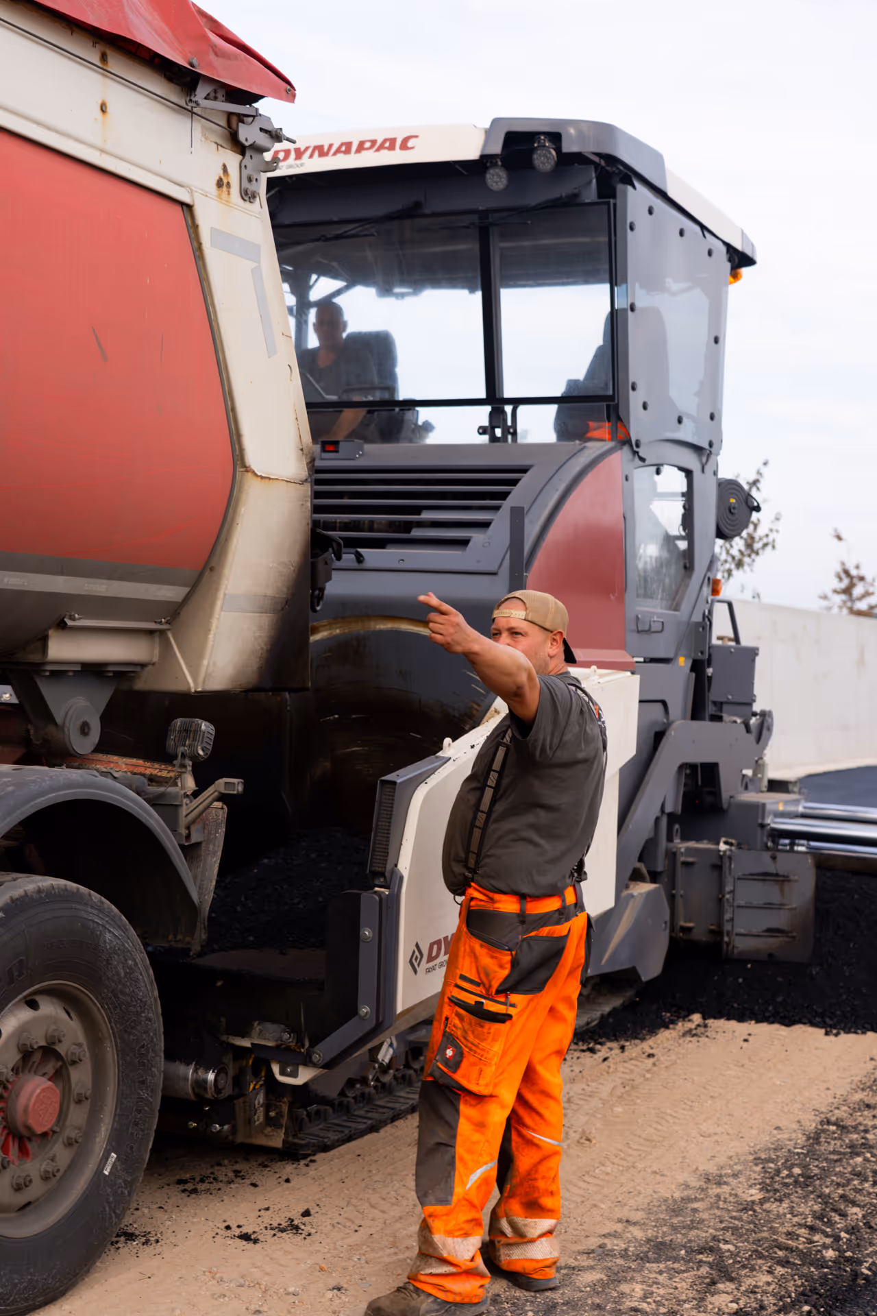 Bauarbeiter in oranger Warnkleidung signalisiert neben Baustellenfahrzeug und Asphaltfertiger.