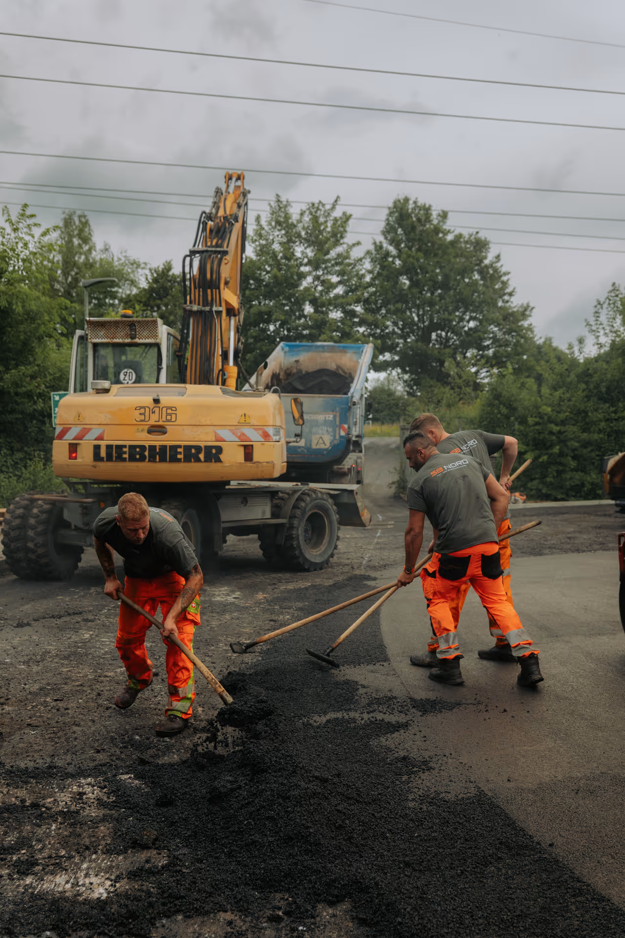 Drei Bauarbeiter mit orangen Hosen verteilen Asphalt auf einer Straße mittels Schaufeln vor einer Baumaschine.