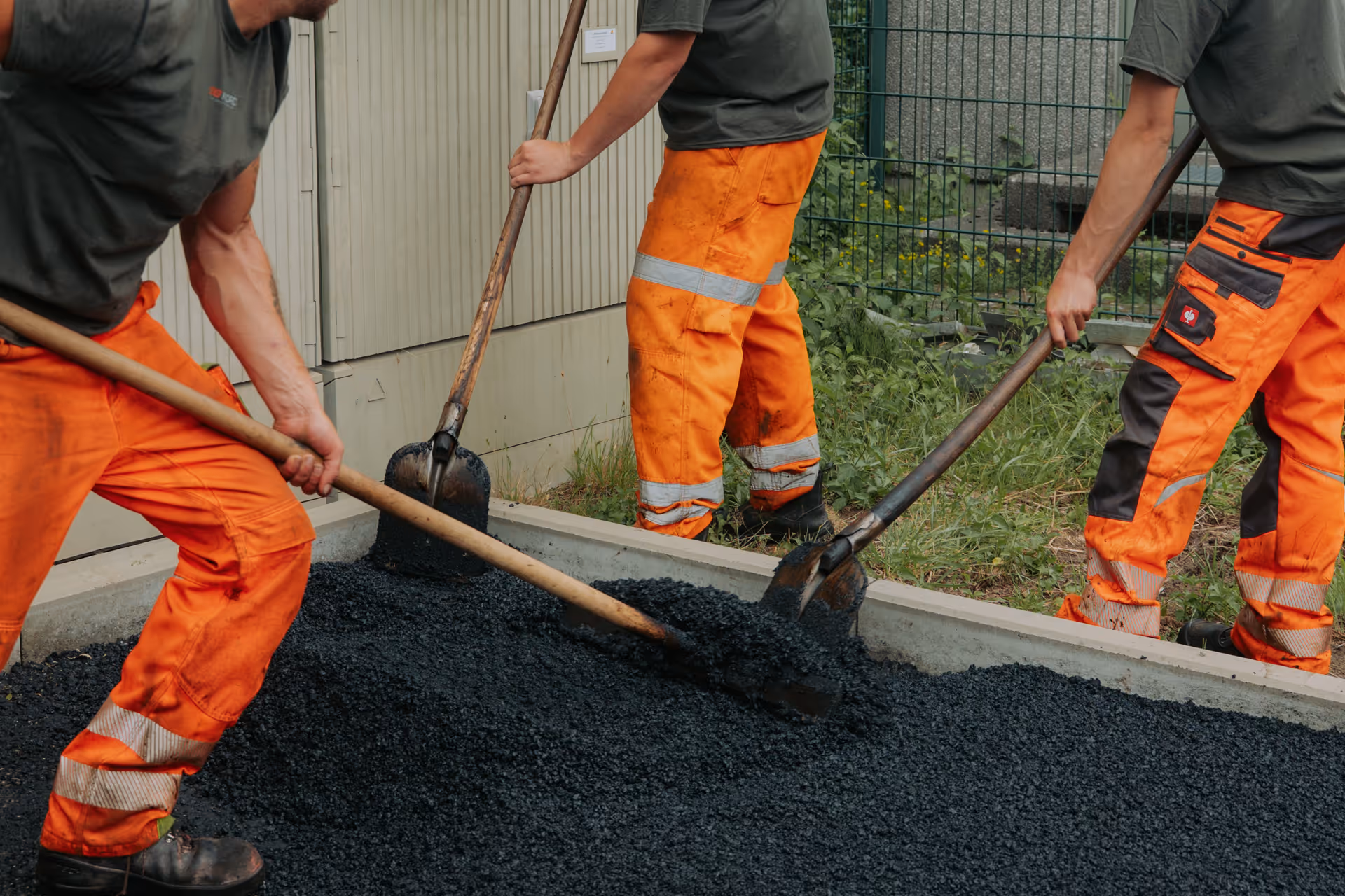 Drei Arbeiter in orangefarbenen Hosen schaufeln Asphalt auf einen Straßenrand.