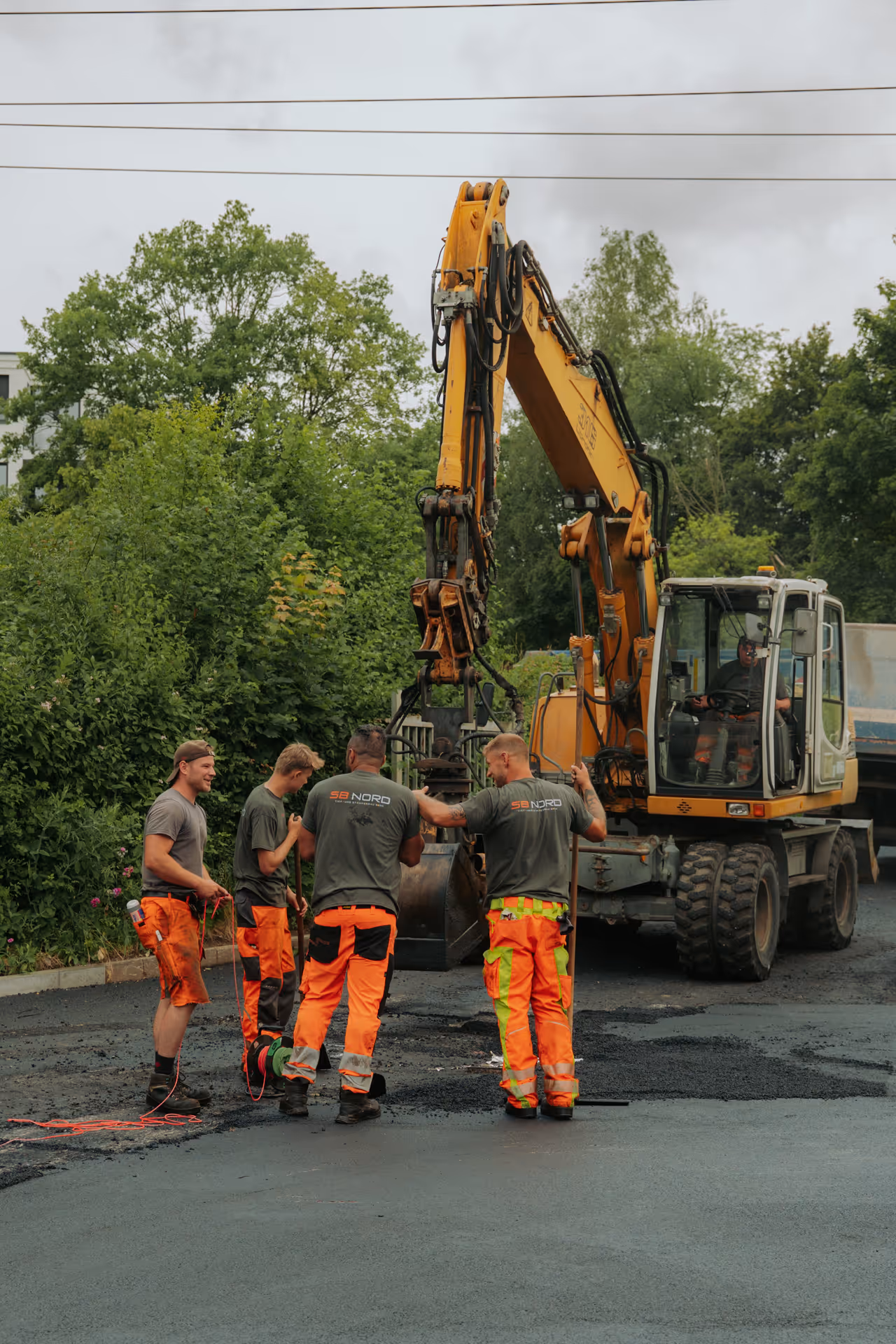 Four construction workers in orange pants working with a large yellow excavator on a road surrounded by greenery.