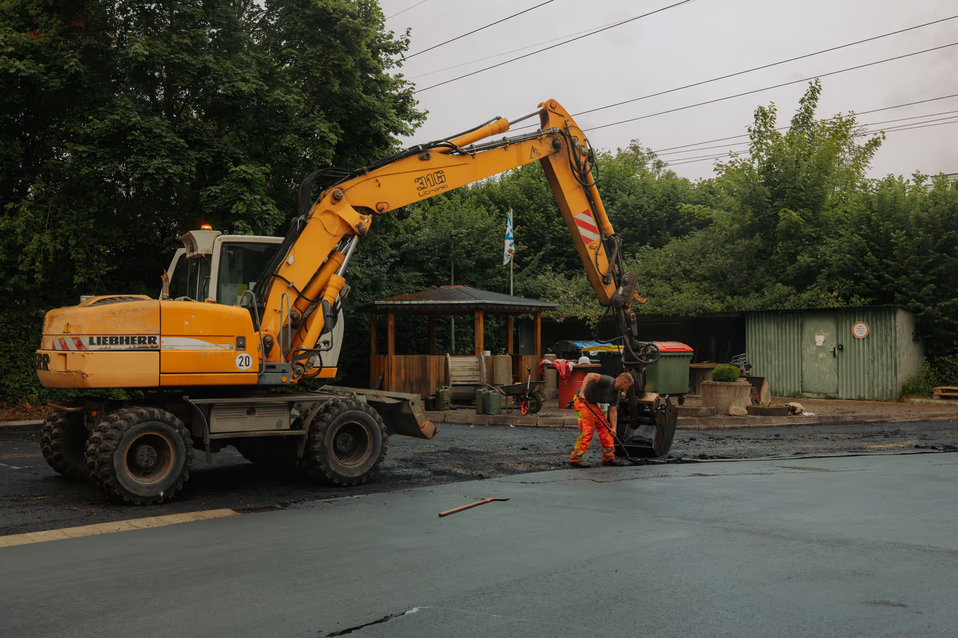Construction worker in orange pants using a tool near a yellow excavator on a paved area surrounded by trees and a small green shed.