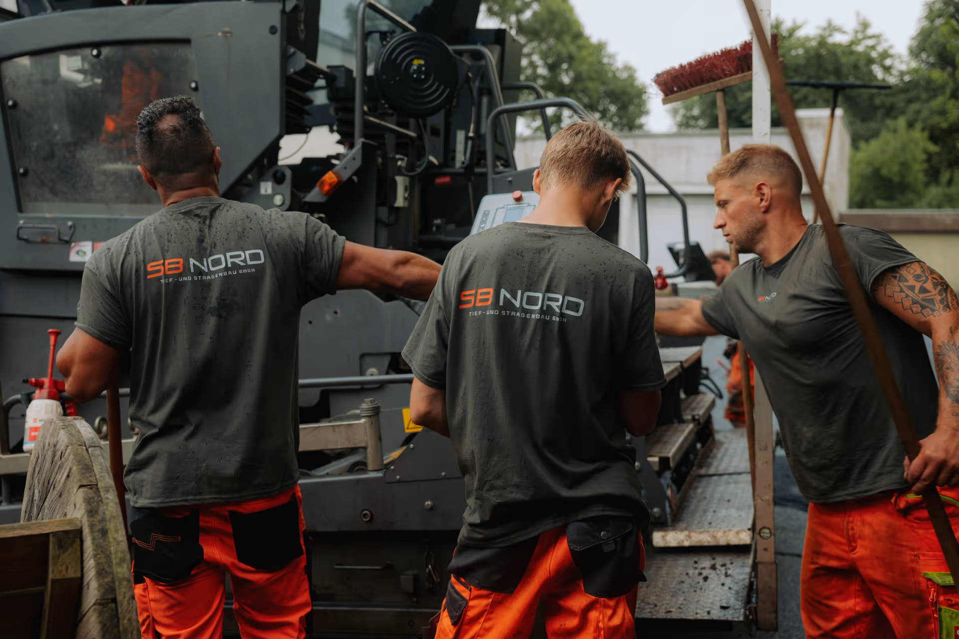 Drei Bauarbeiter in Arbeitskleidung mit SB NORD-Logo auf dem Rücken arbeiten an einer Straßenbaumaschine.