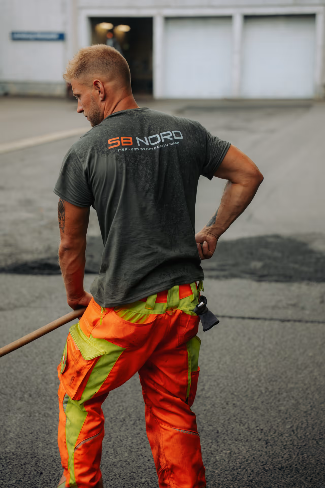 Bauarbeiter mit orange-grüner Arbeitshose und grauem T-Shirt arbeitet mit Schaufel auf Straße.