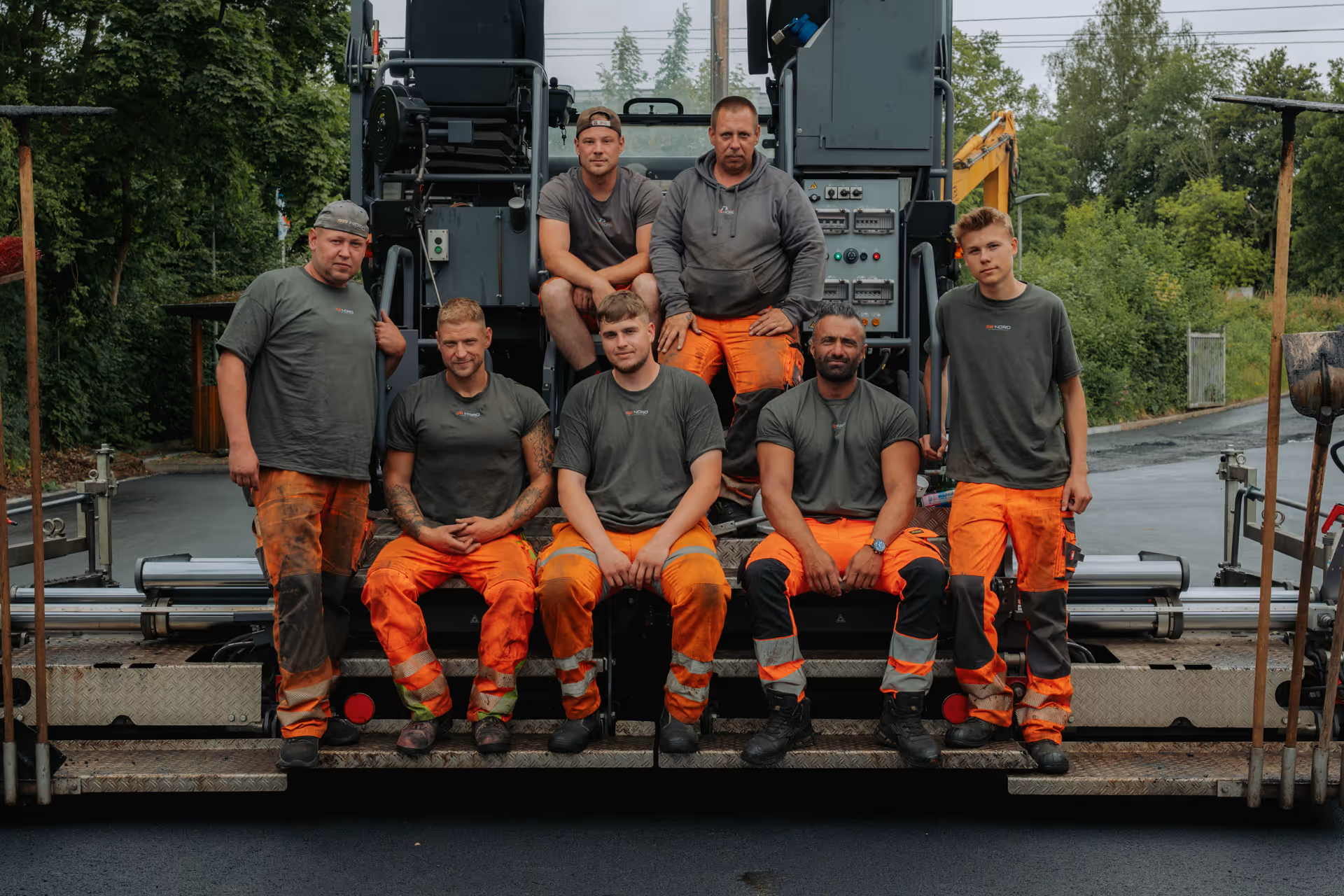 Sieben Bauarbeiter mit orangen Hosen und grauen Shirts sitzen auf einer Maschine im Freien.
