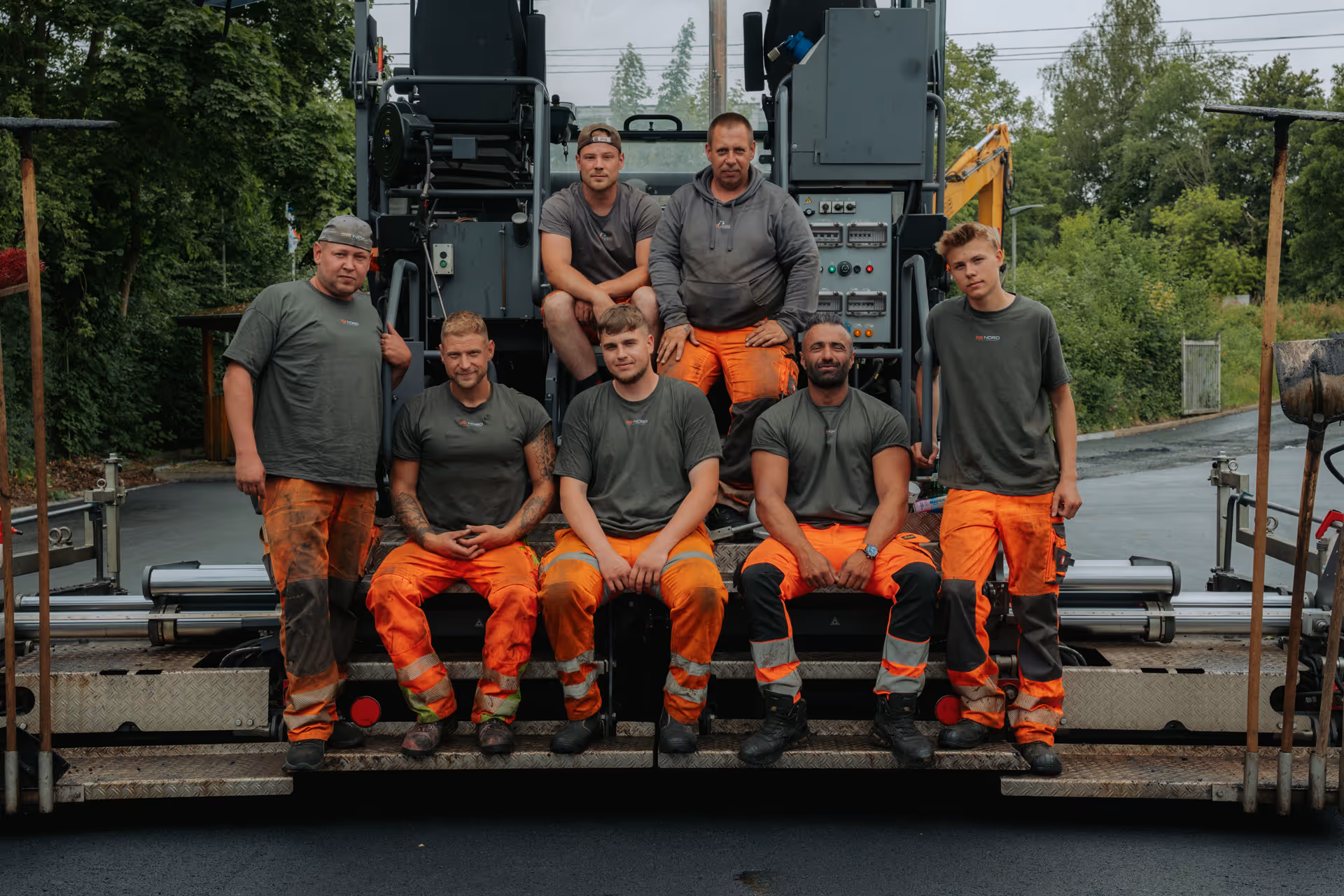 Seven construction workers wearing gray shirts and orange safety pants posing on and in front of a road paving machine on a freshly paved street.