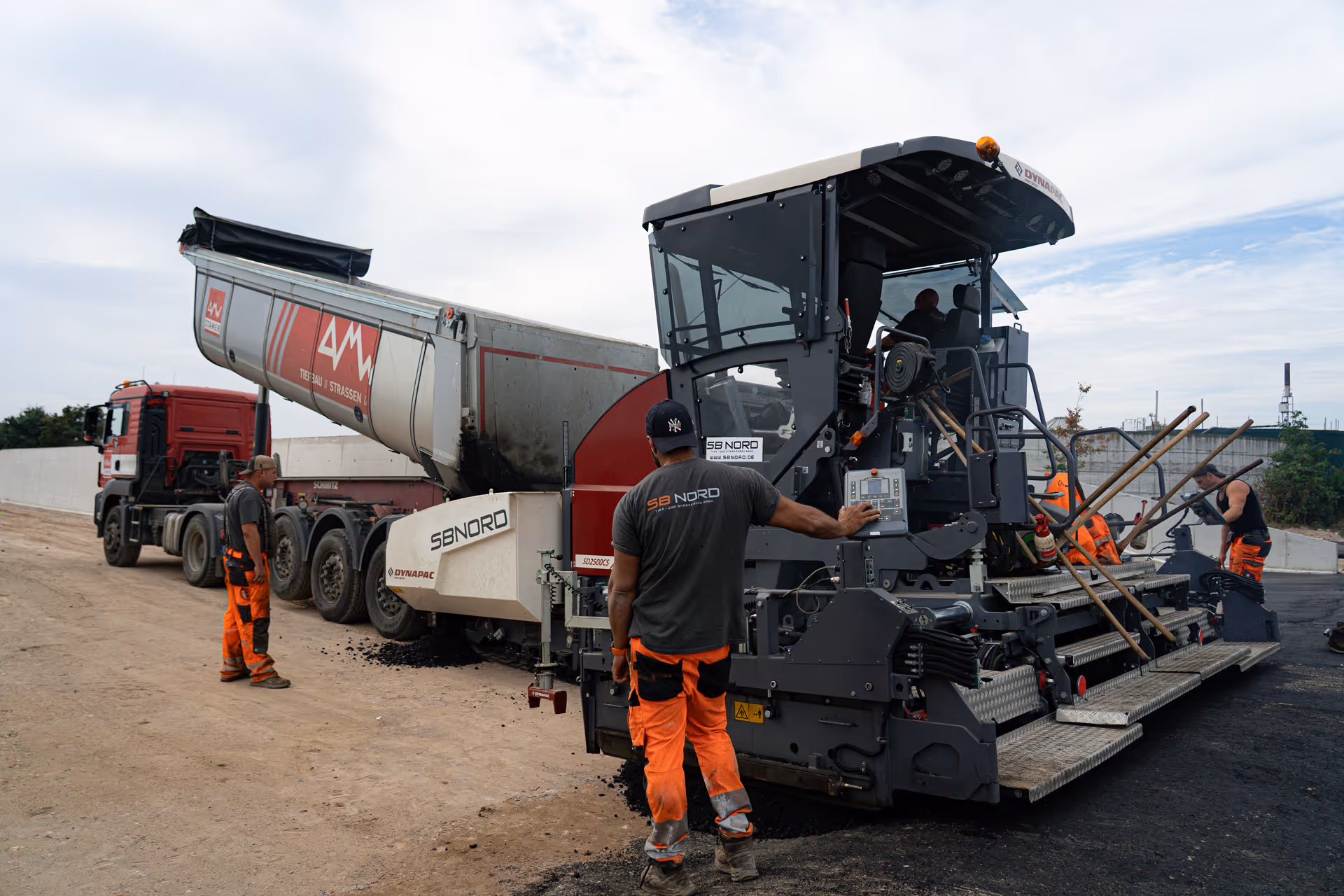 Construction workers operating heavy machinery to lay asphalt on a road beside a large truck.