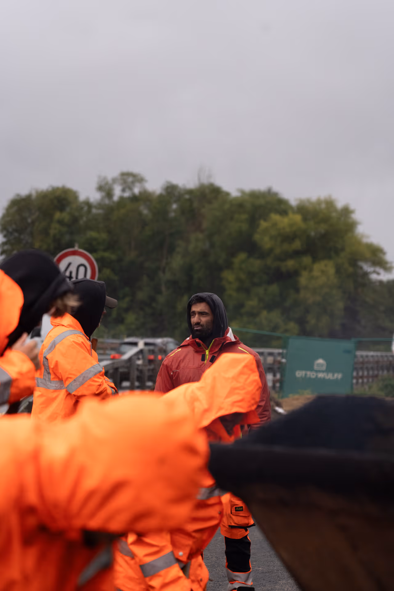 Person in red jacket with hood among workers in orange high-visibility rain gear outdoors near trees.