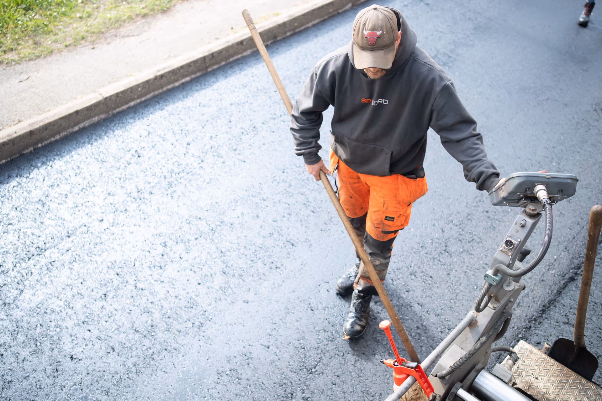 Bauarbeiter mit oranger Arbeitskleidung und grauem Kapuzenpullover glättet frisch asphaltierte Straße mit einem Holzwerkzeug.