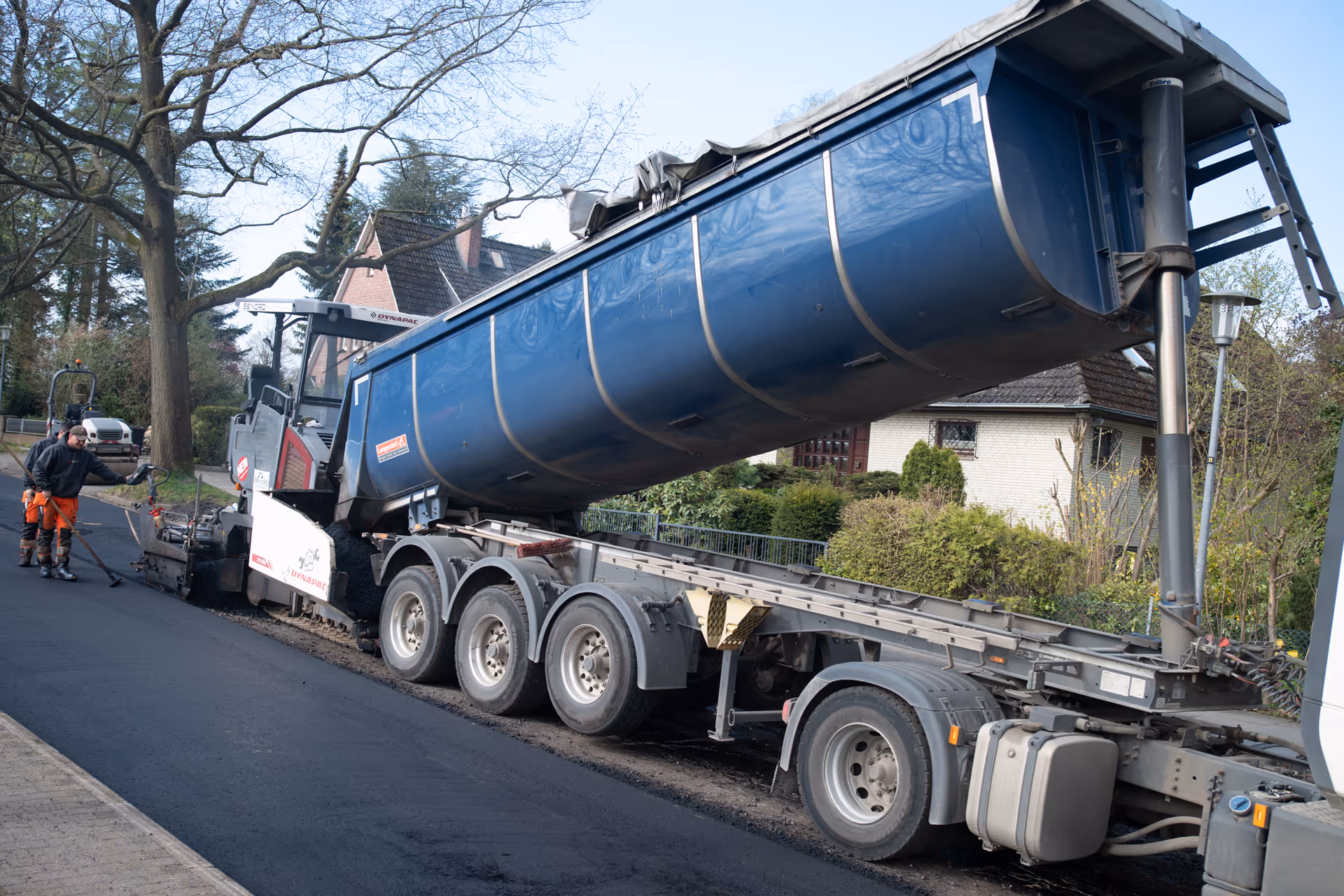 Straßenbauarbeiter arbeiten mit einer Maschine, die Asphalt von einem gekippten blauen LKW ablädt.