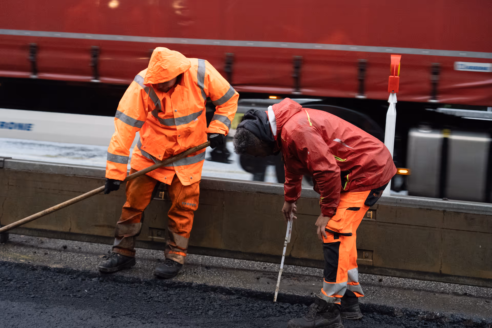 Zwei Bauarbeiter in reflektierender Kleidung reparieren eine Straße bei nassem Wetter vor einem LKW.