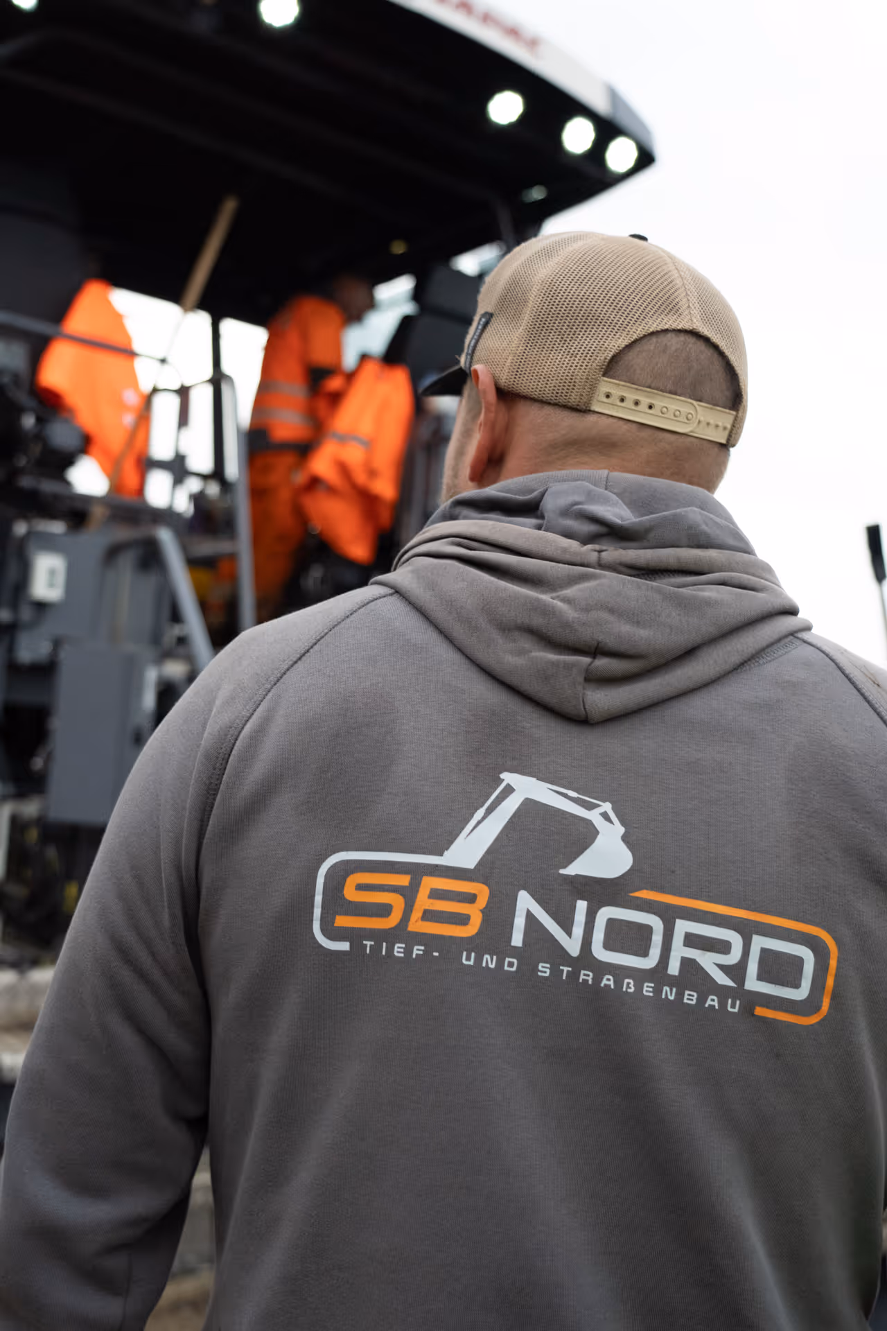 Rückansicht eines Mannes in grauem Hoodie mit SB NORD Logo vor Bauarbeitern in orangefarbener Arbeitskleidung.