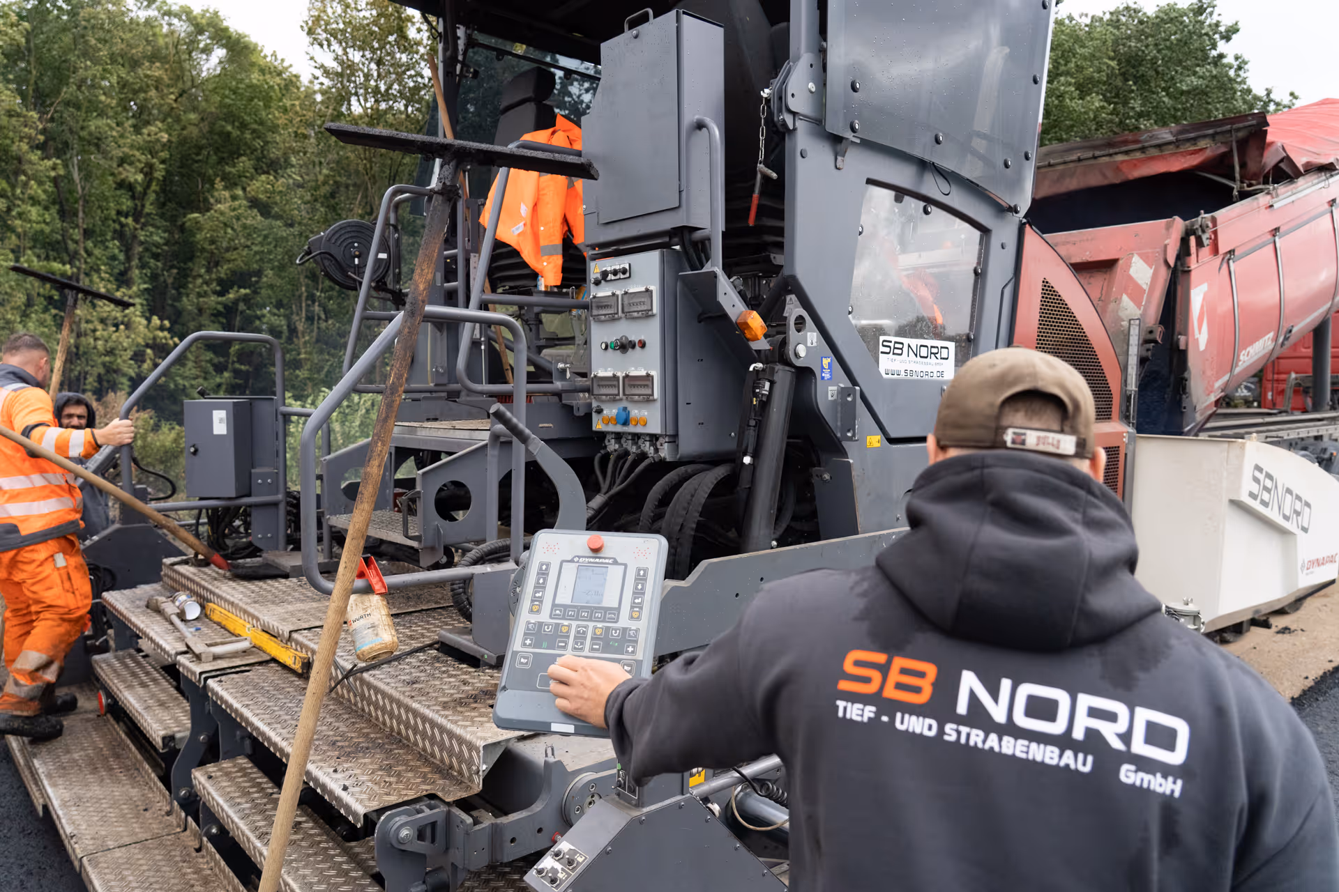 Arbeiter bedienen Straßenbaumaschine mit SB NORD Logo und orangener Arbeitskleidung im Freien.