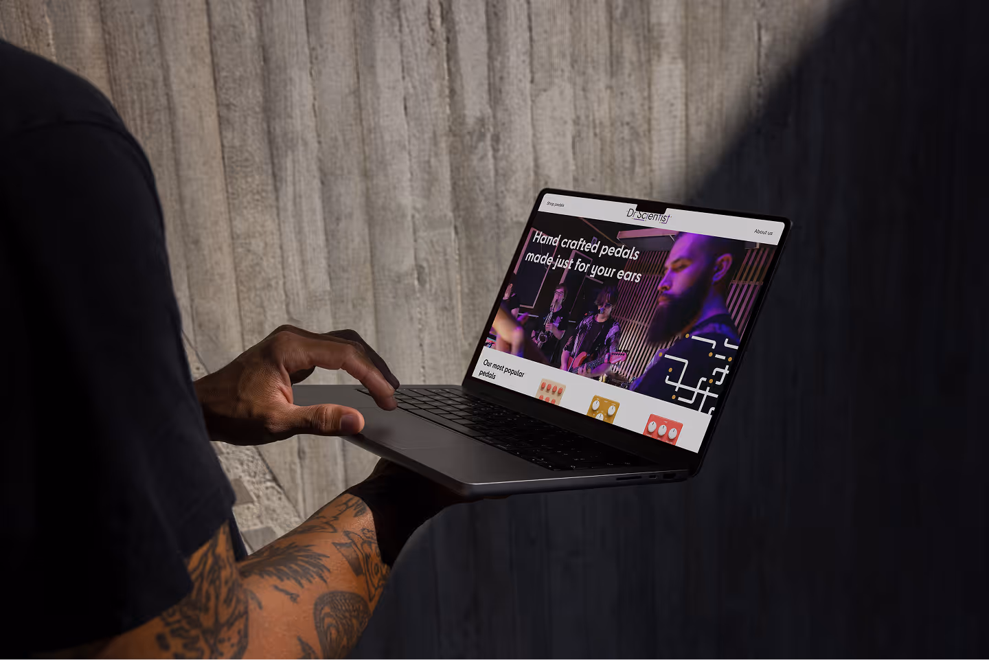 Person with a tattooed arm holding a laptop showing a website for handcrafted pedals.