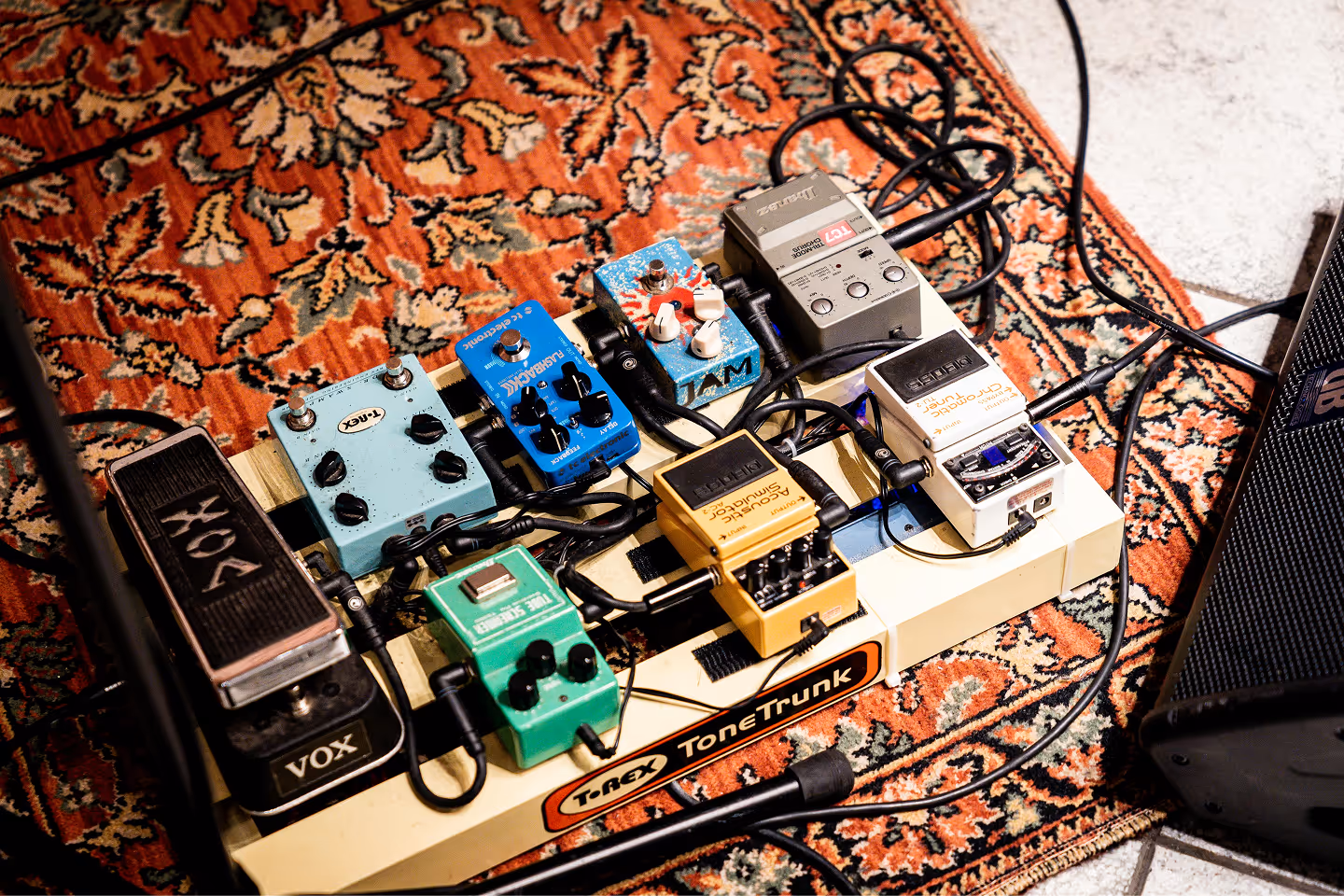 Guitar effects pedalboard with Vox wah, delay, chorus, fuzz, and other pedals on a patterned rug.