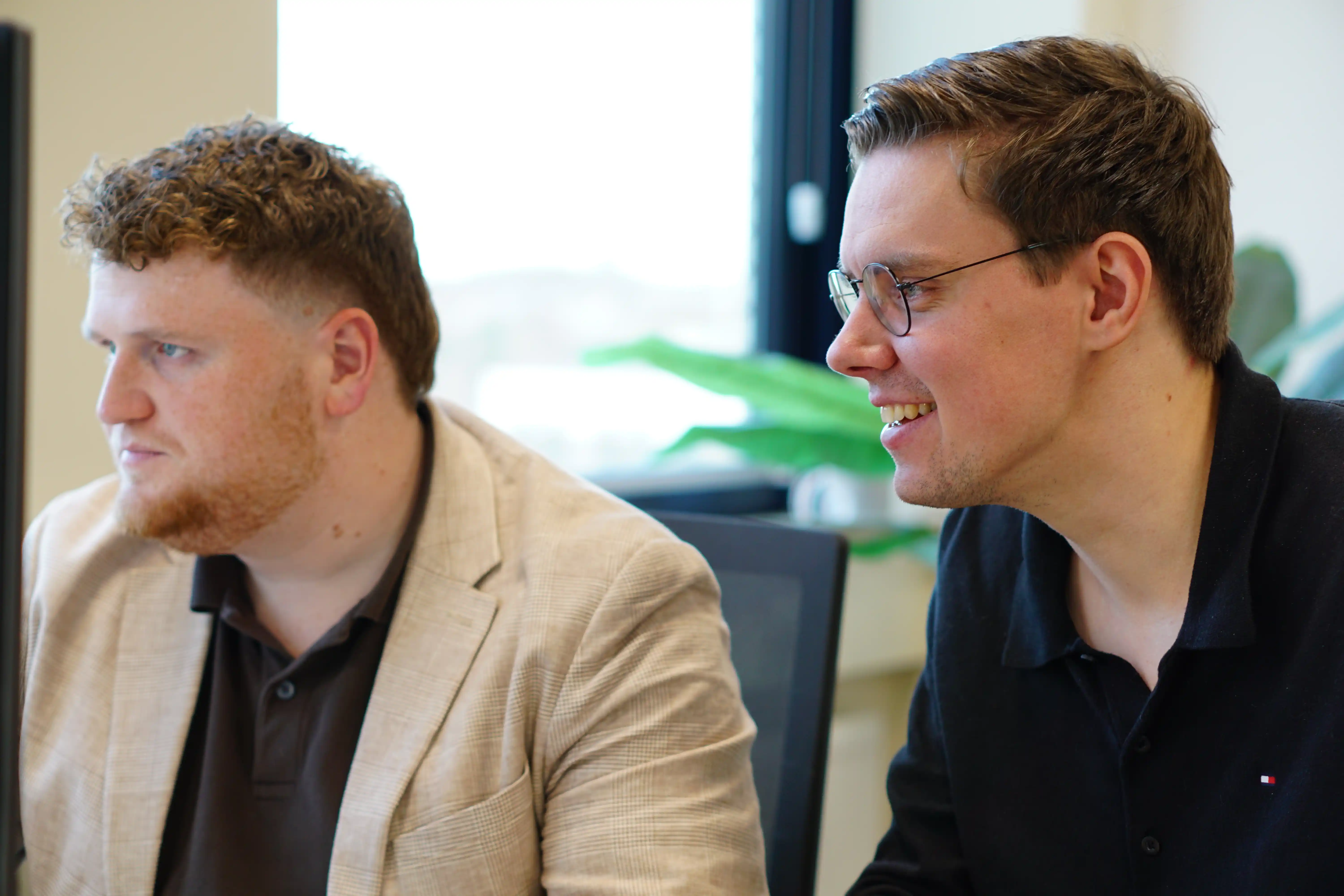 Twee mannen zitten naast elkaar en kijken naar iets links buiten beeld met een glimlach en ernstig gezicht.
