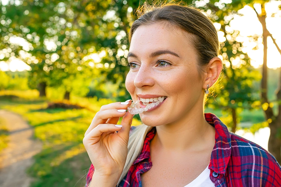 Smiling woman outdoors in plaid shirt putting clear dental aligner in her mouth at sunset.