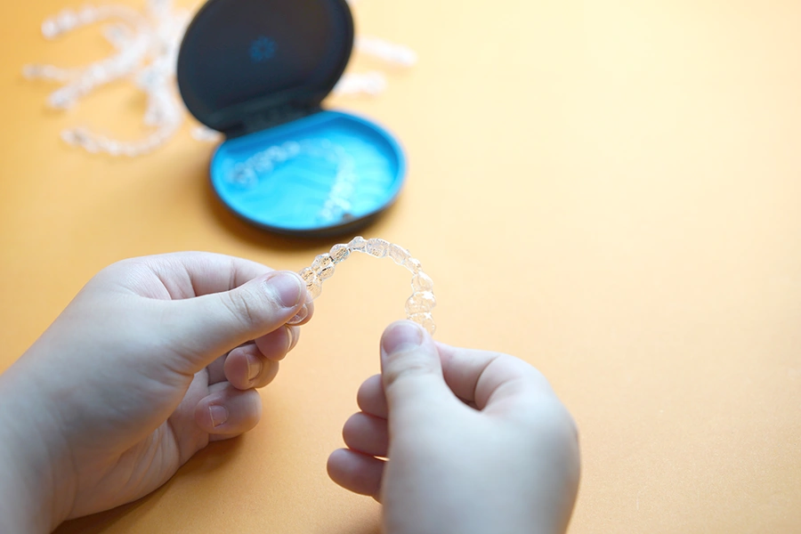Hands holding a clear dental aligner with a blue case in the background on a yellow surface.
