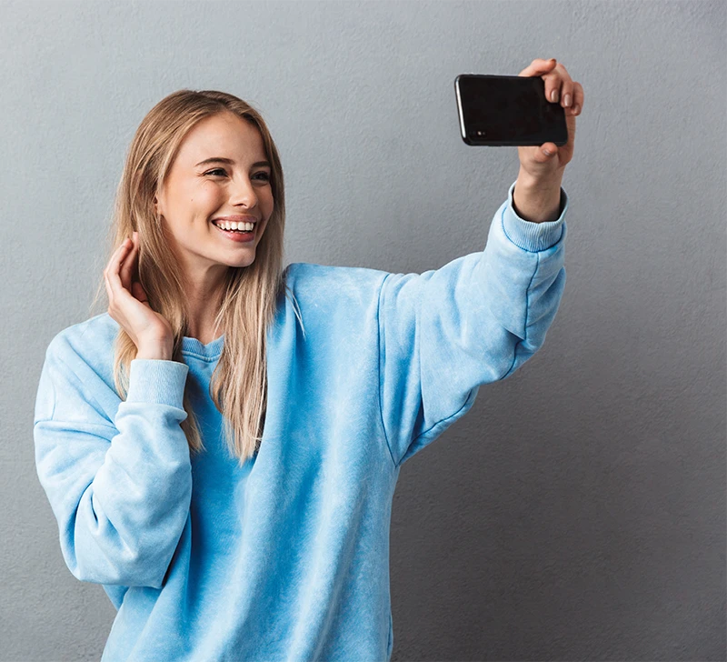 Smiling woman in blue sweatshirt taking selfie with smartphone against gray wall.
