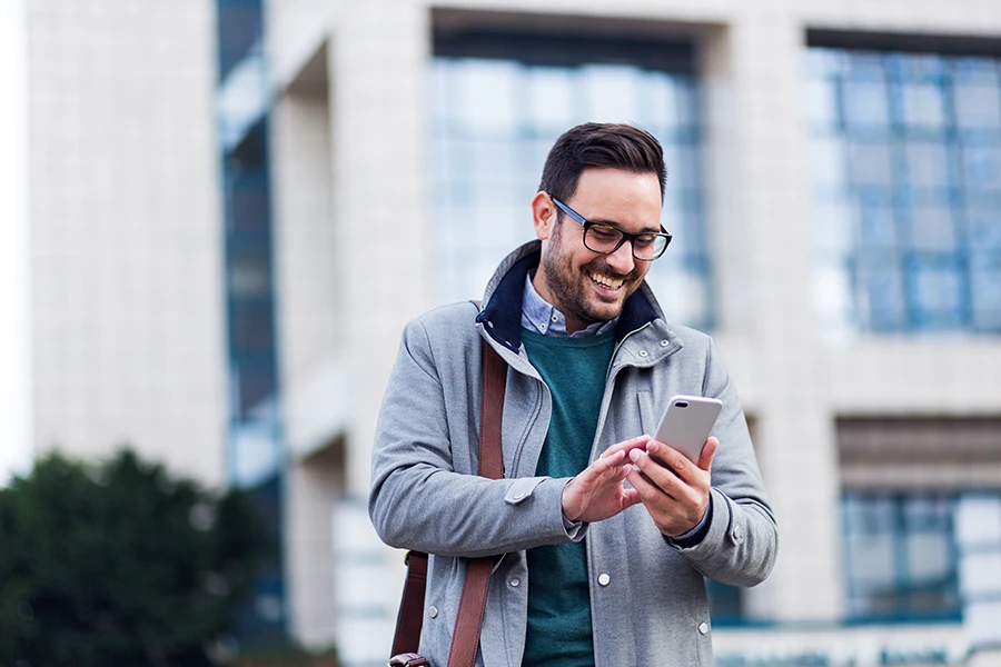 Smiling man wearing glasses and coat using a smartphone outside an office building.