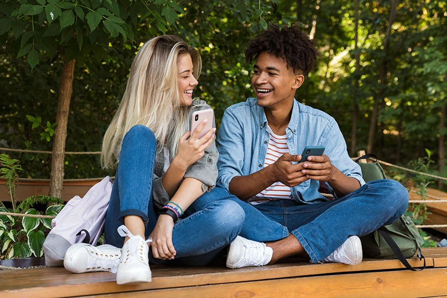 Two young friends sitting outdoors on wooden bench, smiling and holding smartphones.
