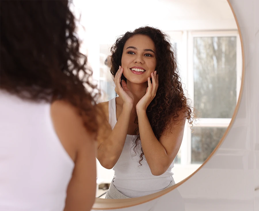 Smiling woman with curly hair looking at her reflection in a round bathroom mirror.