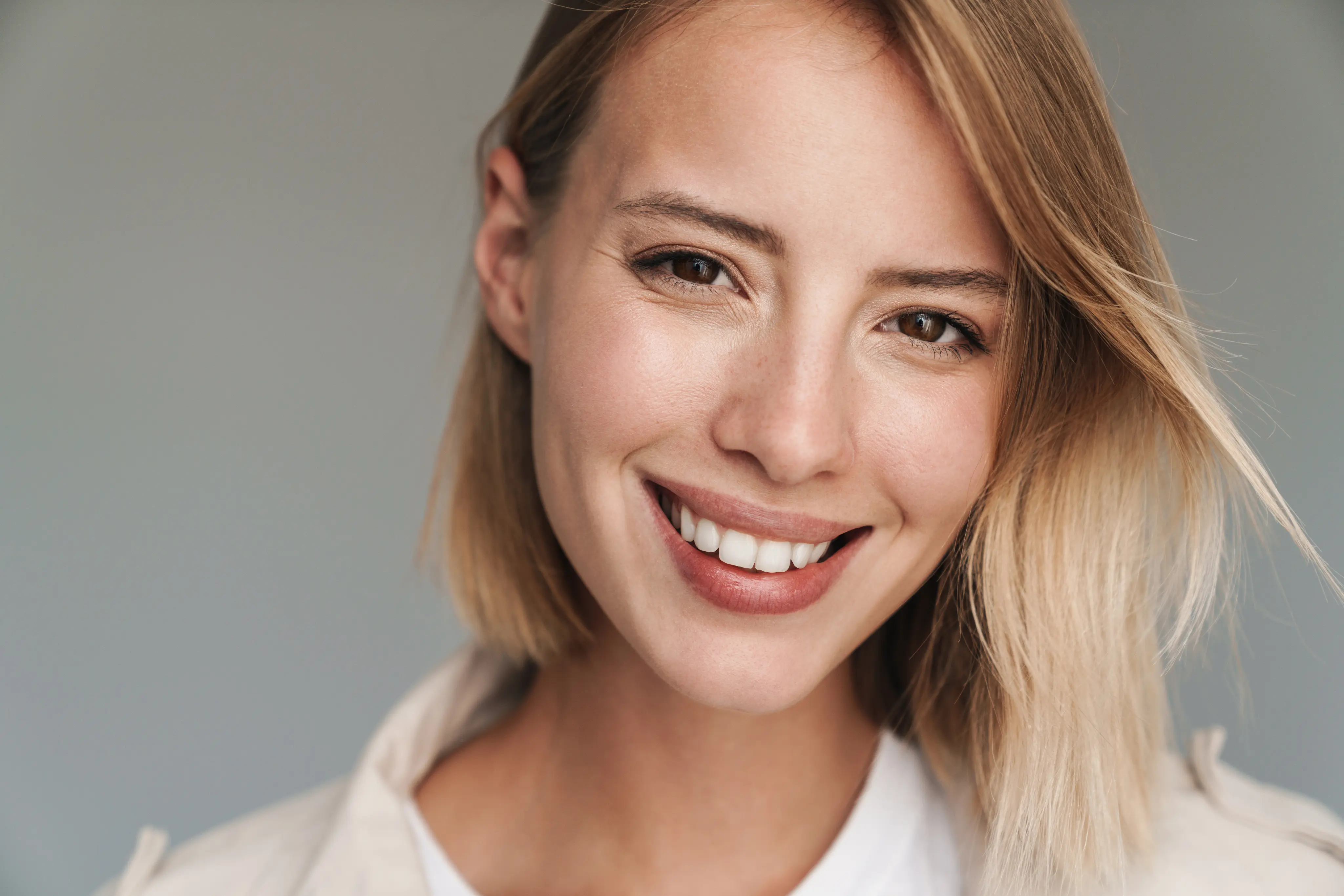 Close-up of a smiling young woman with short blond hair and brown eyes against a gray background.