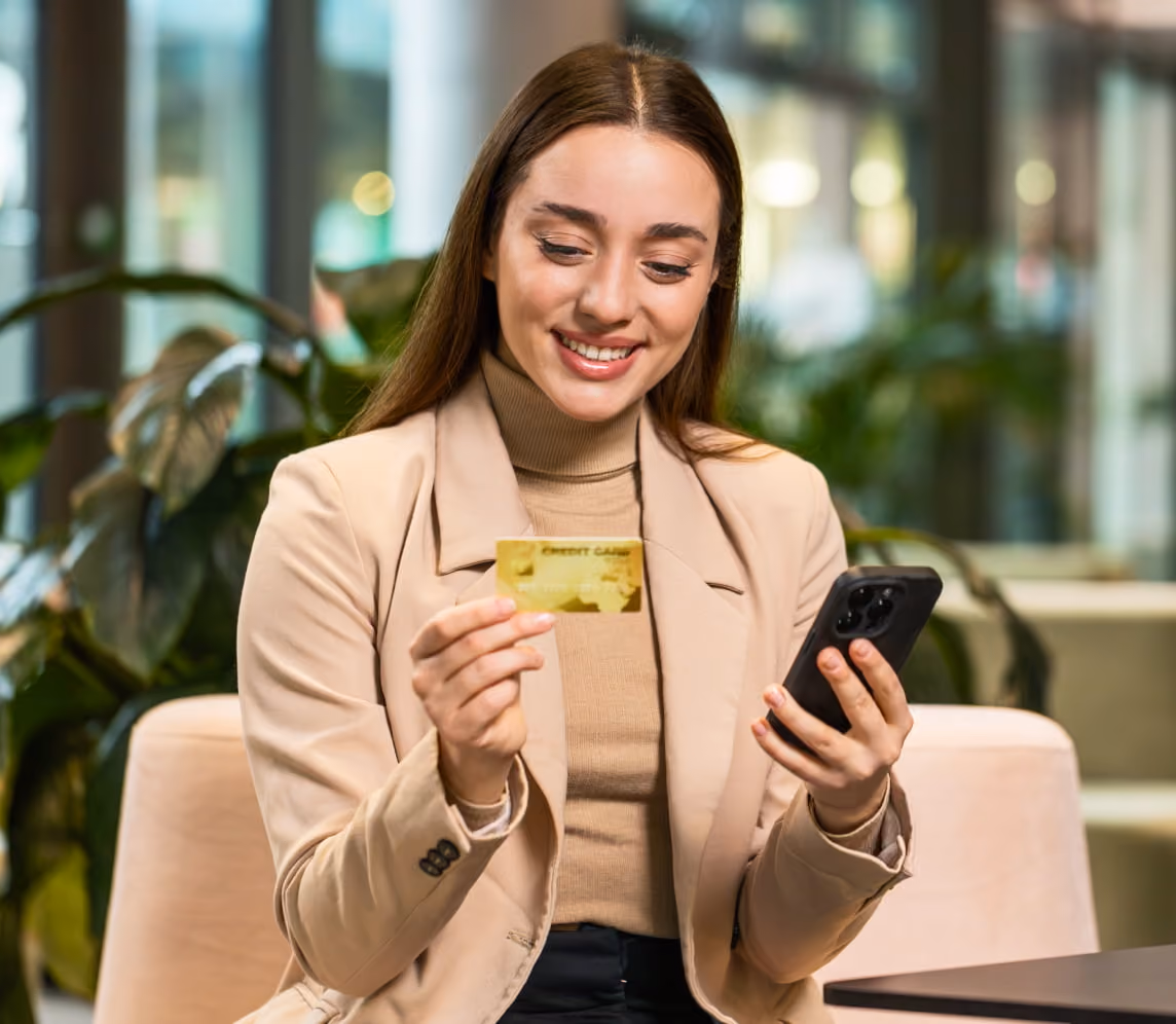 Smiling woman holding a credit card in one hand and a smartphone in the other indoors.
