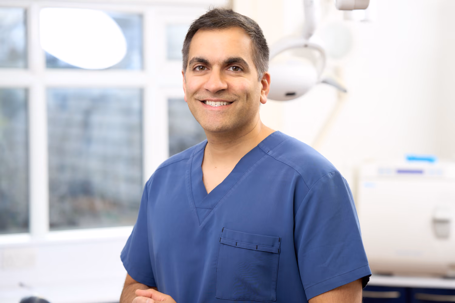 Smiling male healthcare professional wearing blue scrubs in a bright medical office.