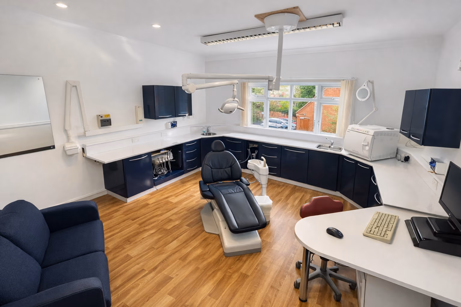Modern dental office with chair, dental tools, desk with computer, and large window overlooking brick buildings.