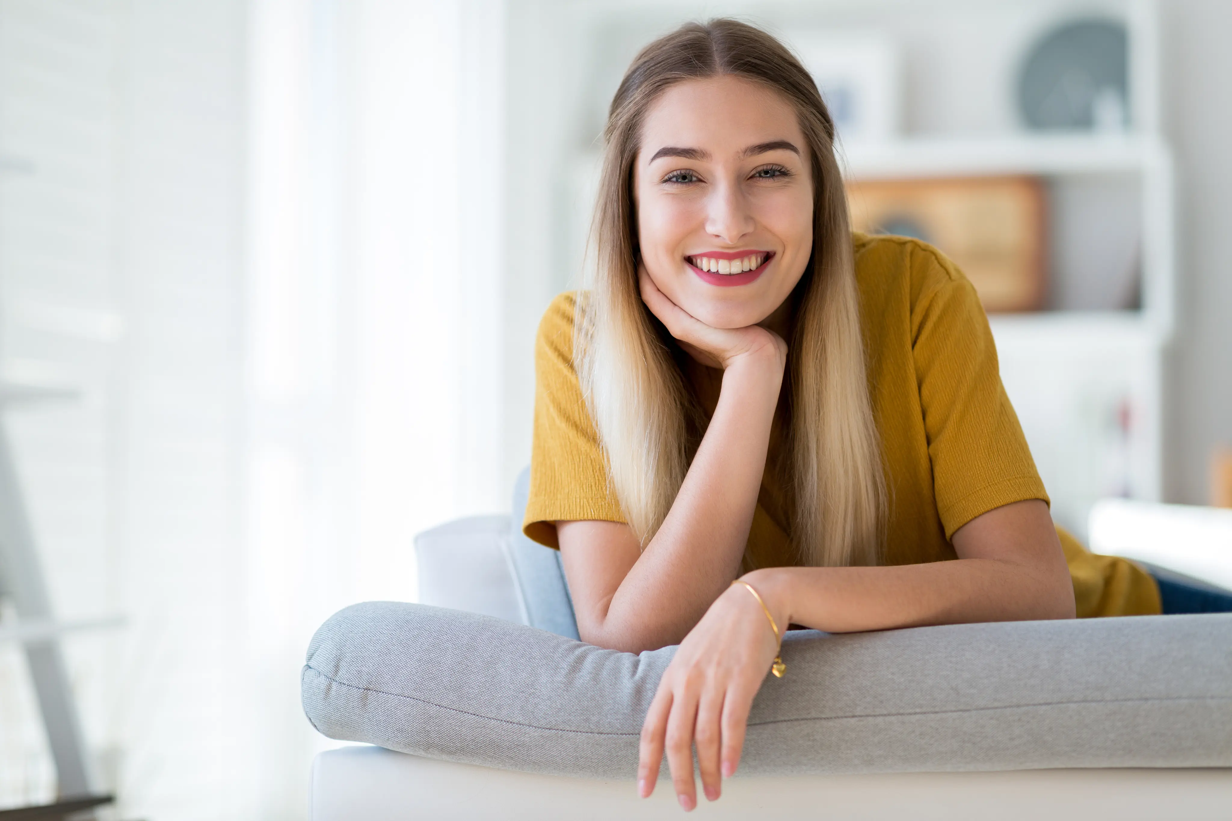 Smiling young woman with long blonde hair resting on a gray cushion in a bright living room.