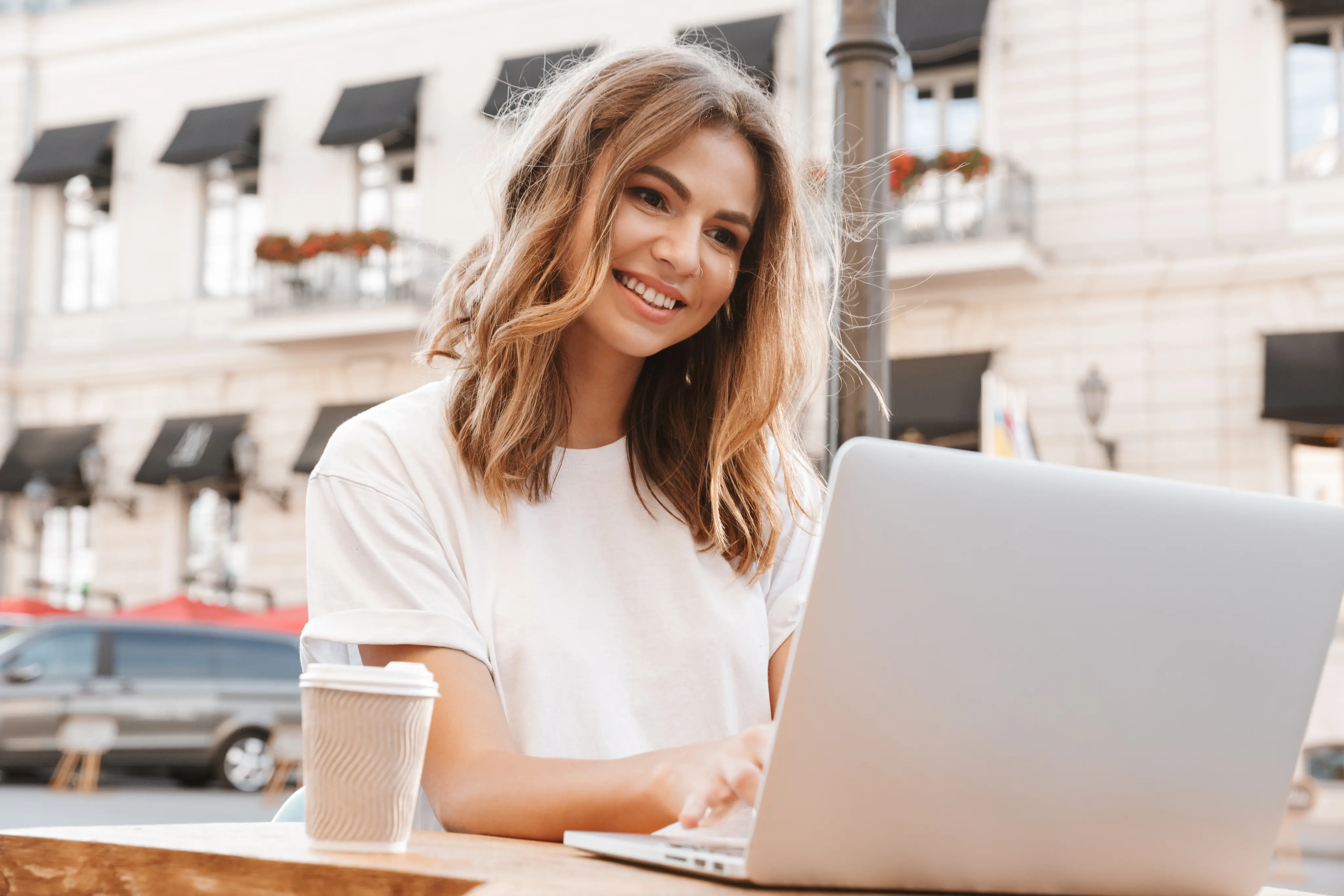 Smiling young woman using a laptop outdoors with coffee cup on the table in an urban setting.