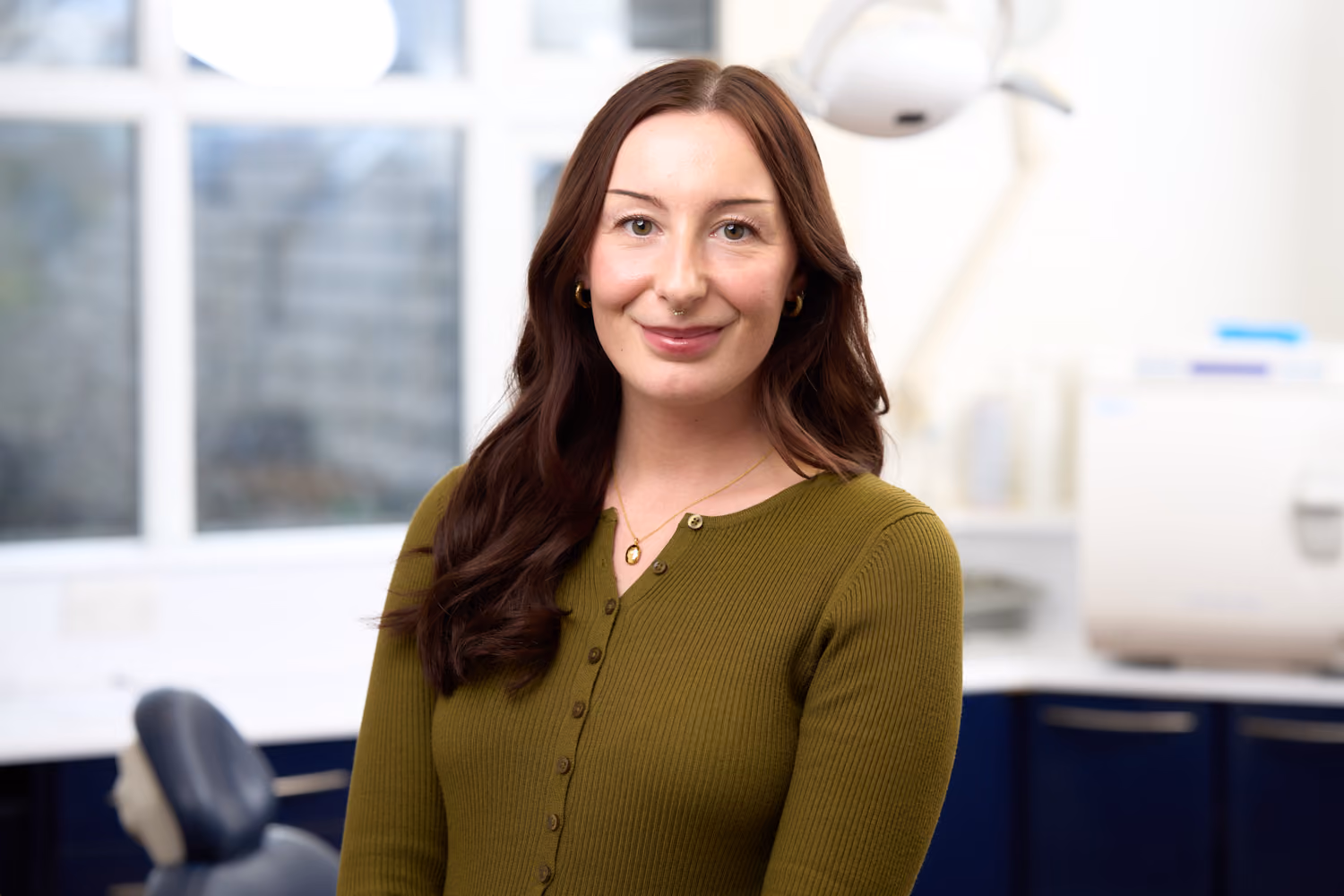 Smiling woman with long brown hair wearing a green top in a bright dental office.