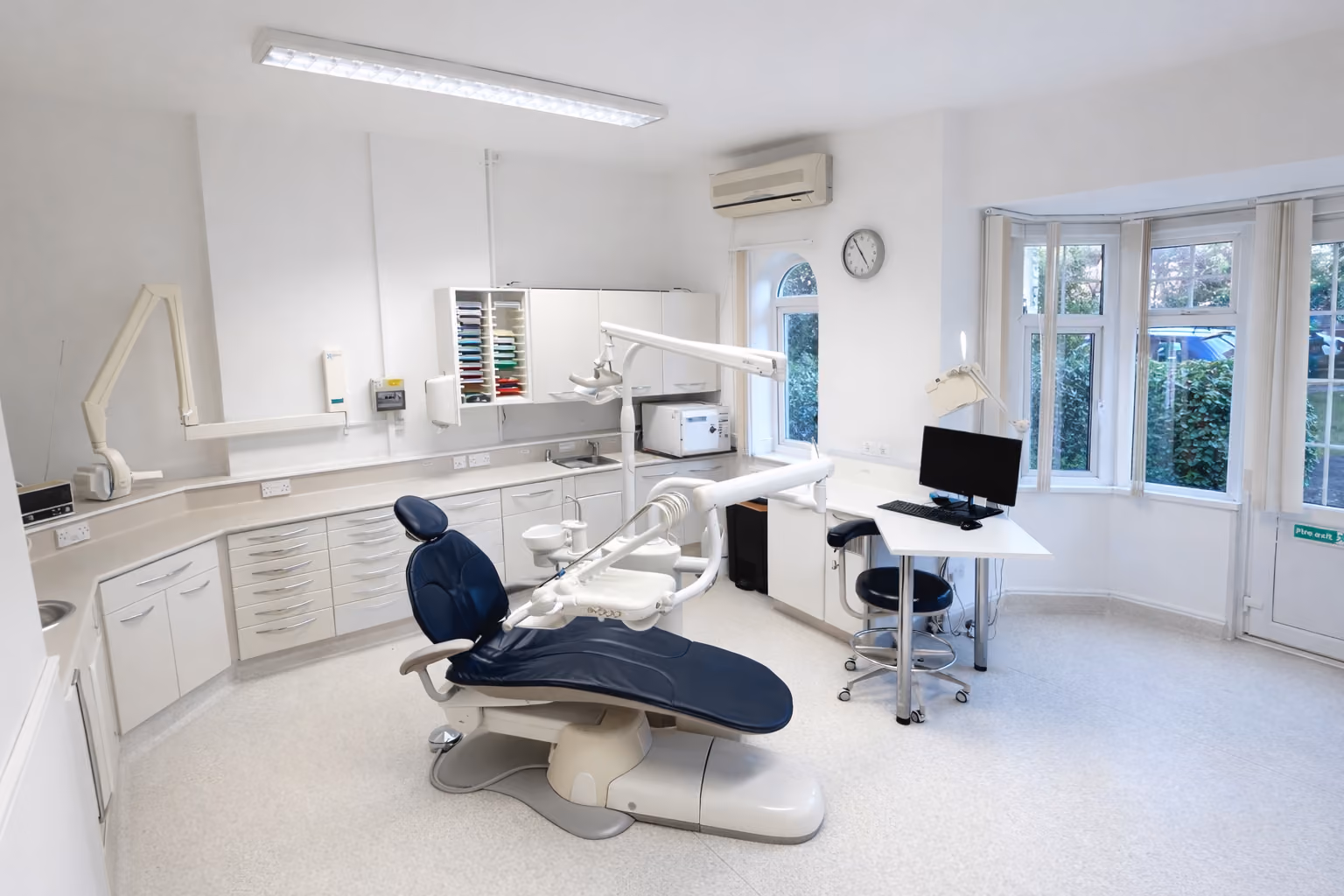 Modern dental clinic with chair, white cabinets, computer, and large windows overlooking greenery.
