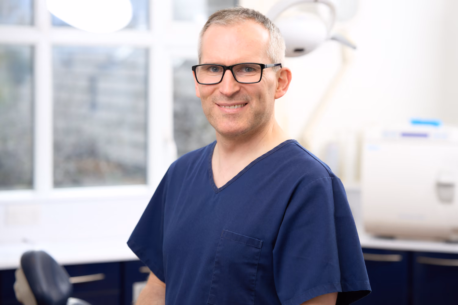 Smiling man in blue scrubs and glasses in a bright dental or medical office.