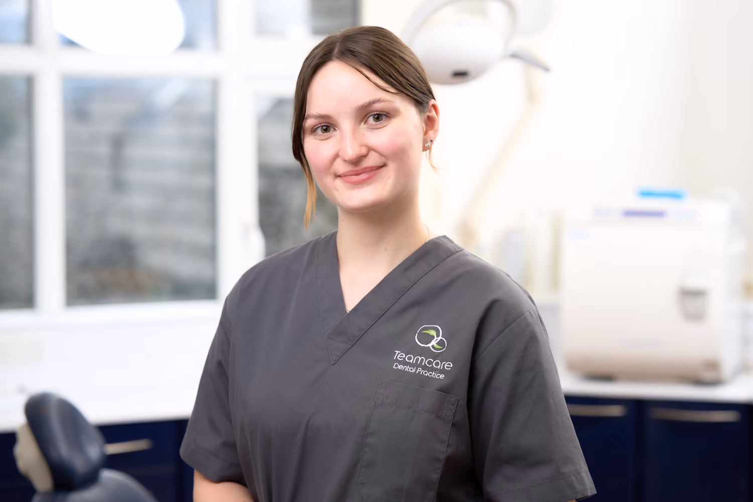 Smiling female dental professional wearing gray scrubs in a dental clinic with equipment in background.