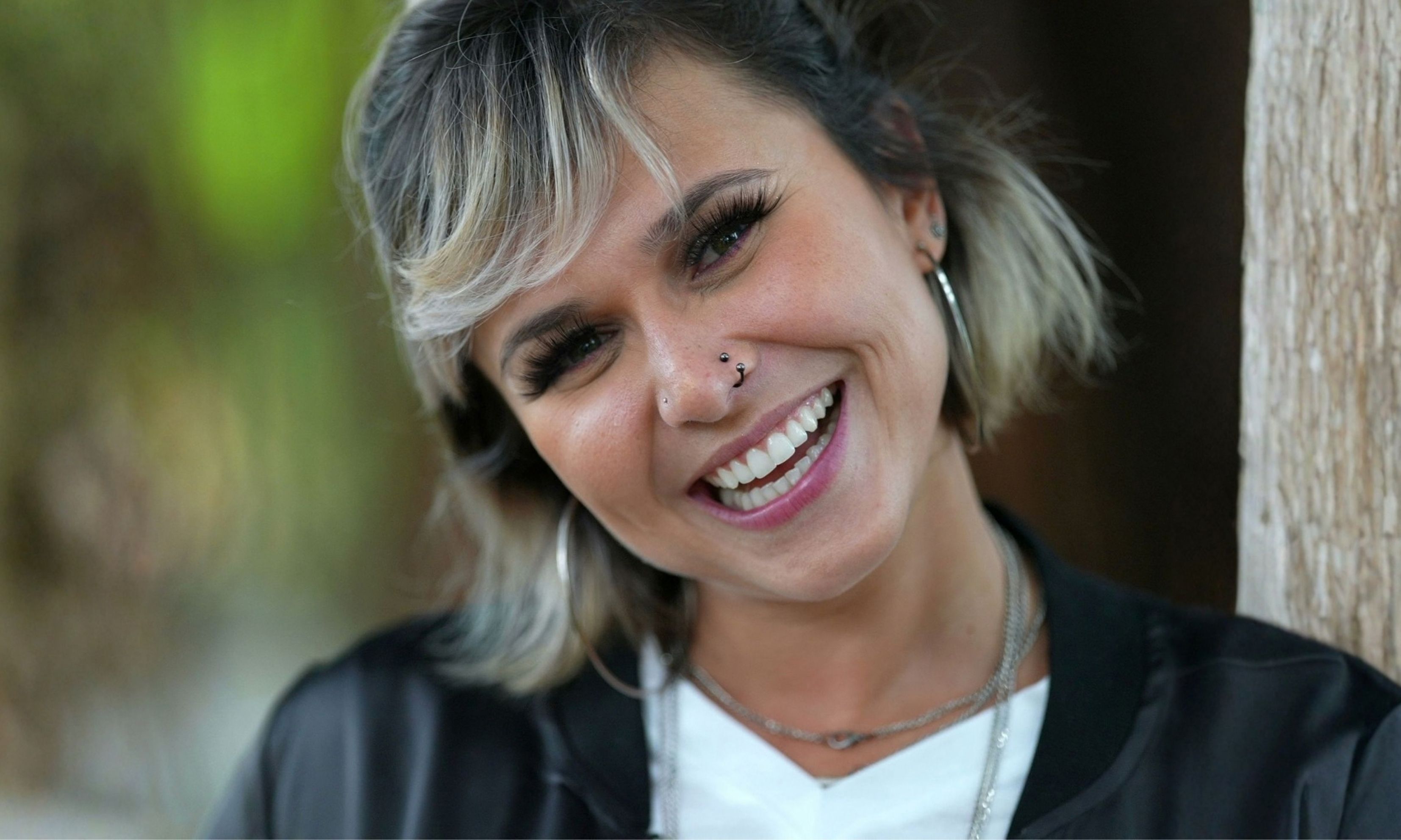 Close-up of a smiling woman with short, blond-highlighted hair, hoop earrings, nose piercings, and layered necklaces, wearing a black jacket over a white top.