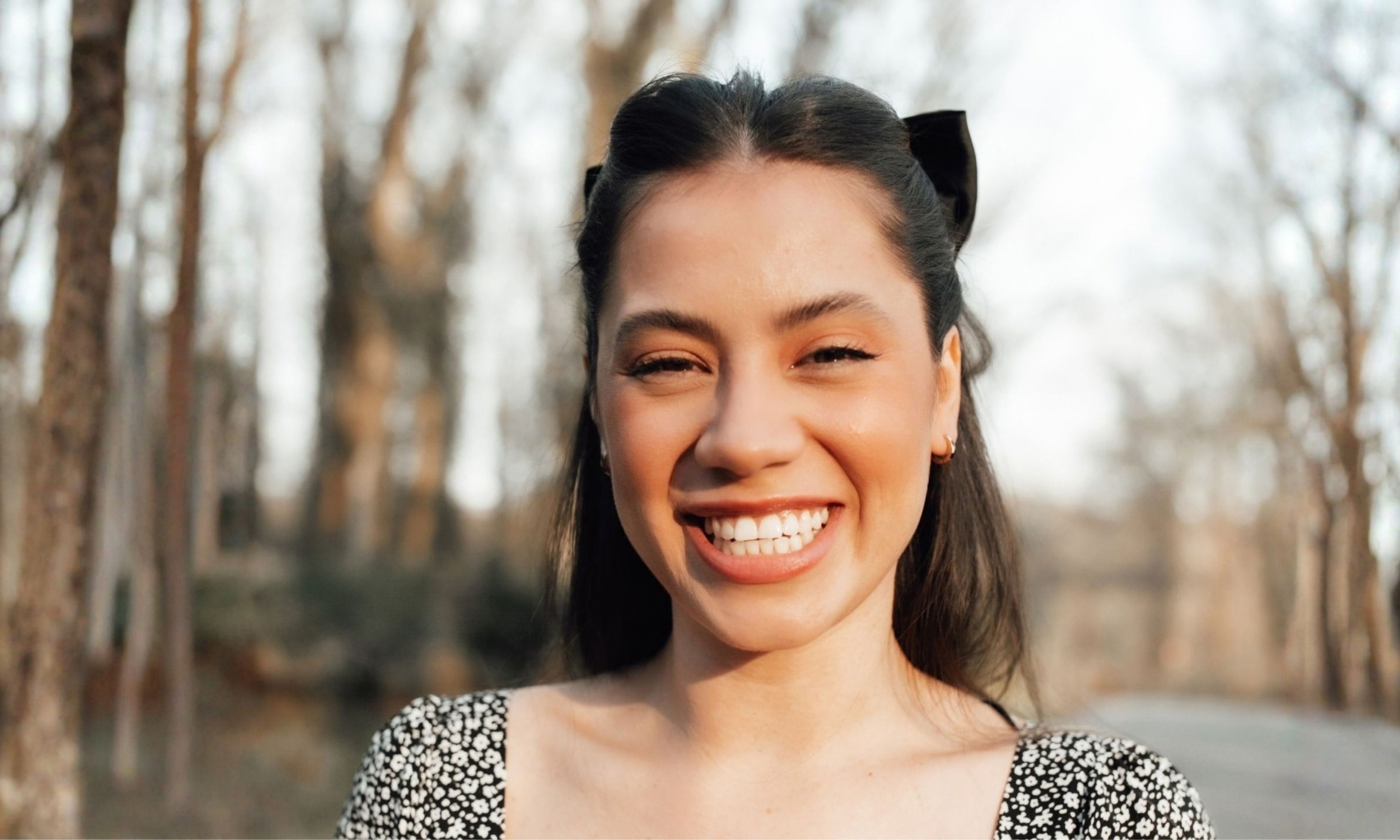 Smiling young woman with long dark hair wearing a black and white floral top, standing outdoors with blurred trees in the background.