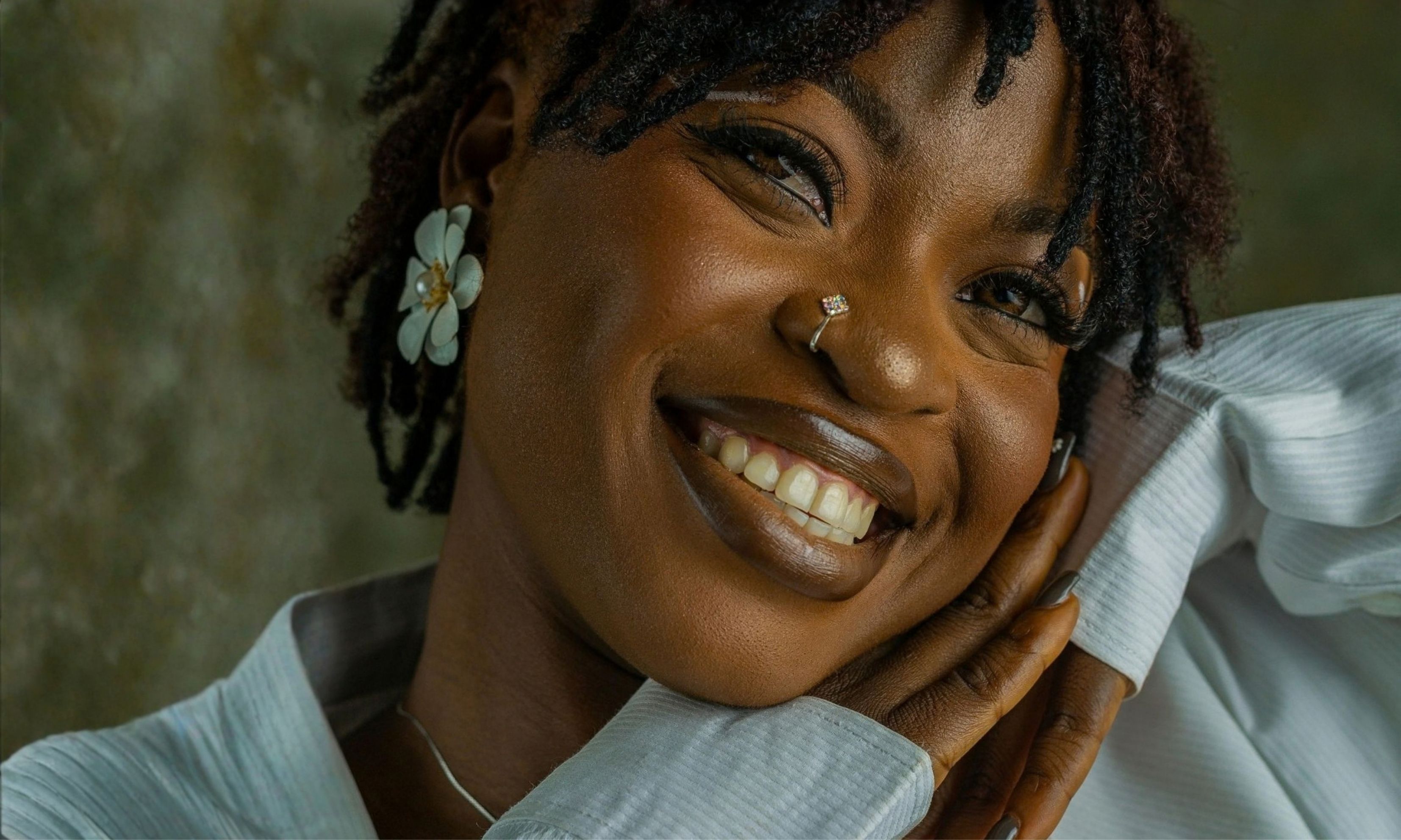 Close-up of a smiling woman with dark skin, wearing a white textured top, a flower-shaped earring, a nose ring, and posing with hands near her face.