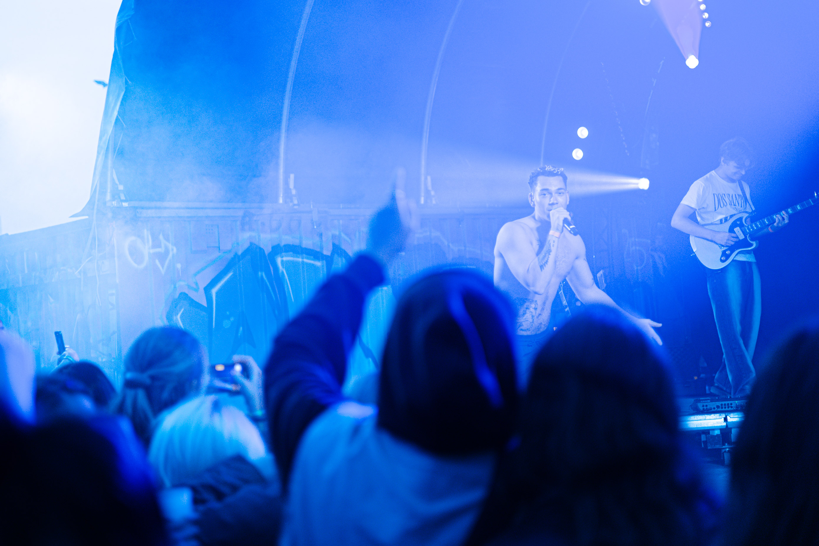 Shirtless male singer with tattoos holding a microphone on stage with guitarist, in blue lighting.