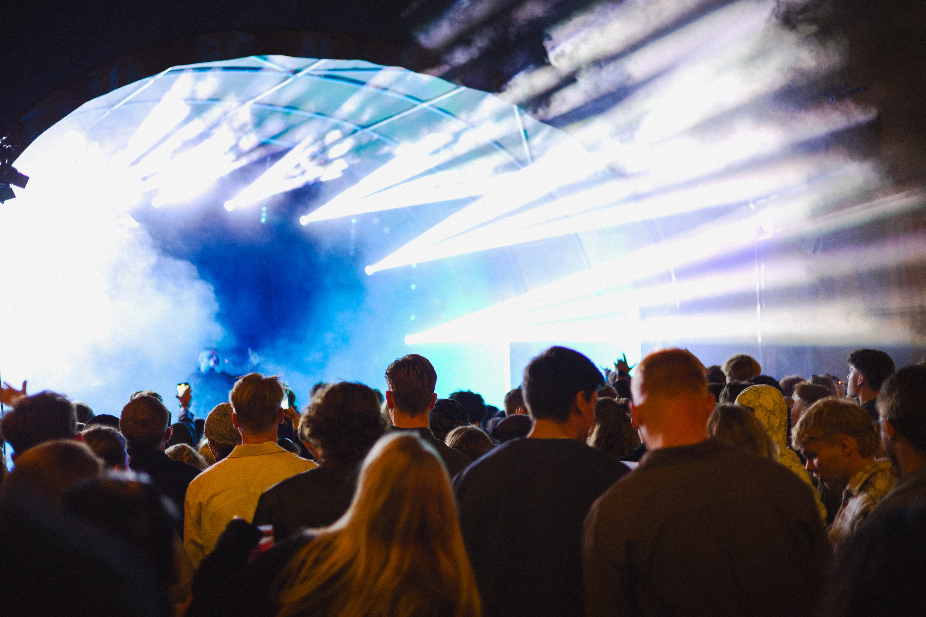Crowd of people watching a concert with bright white and blue stage lights and smoke effects.