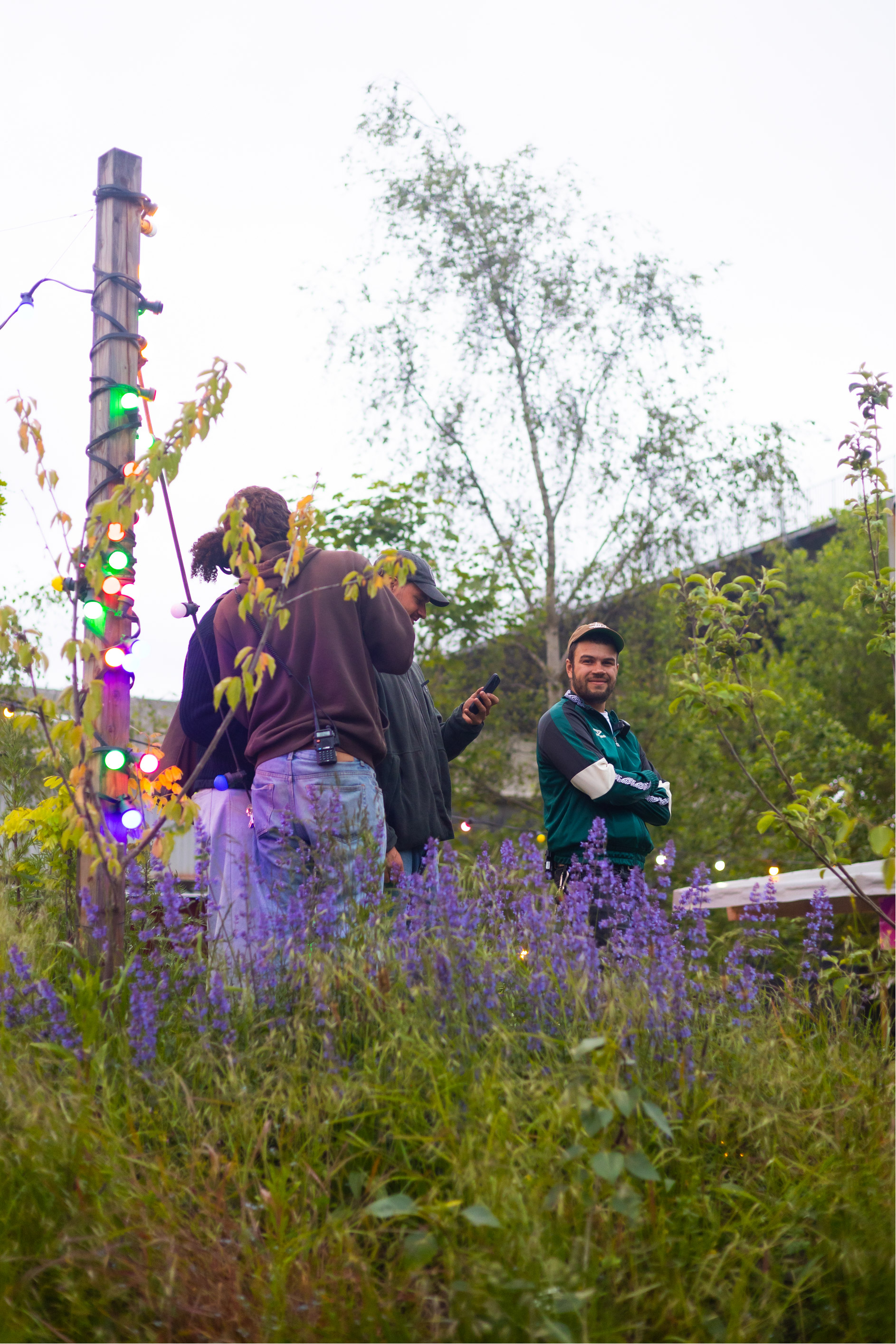 Group of people standing near a wooden post wrapped with colorful string lights in a garden with purple flowers.