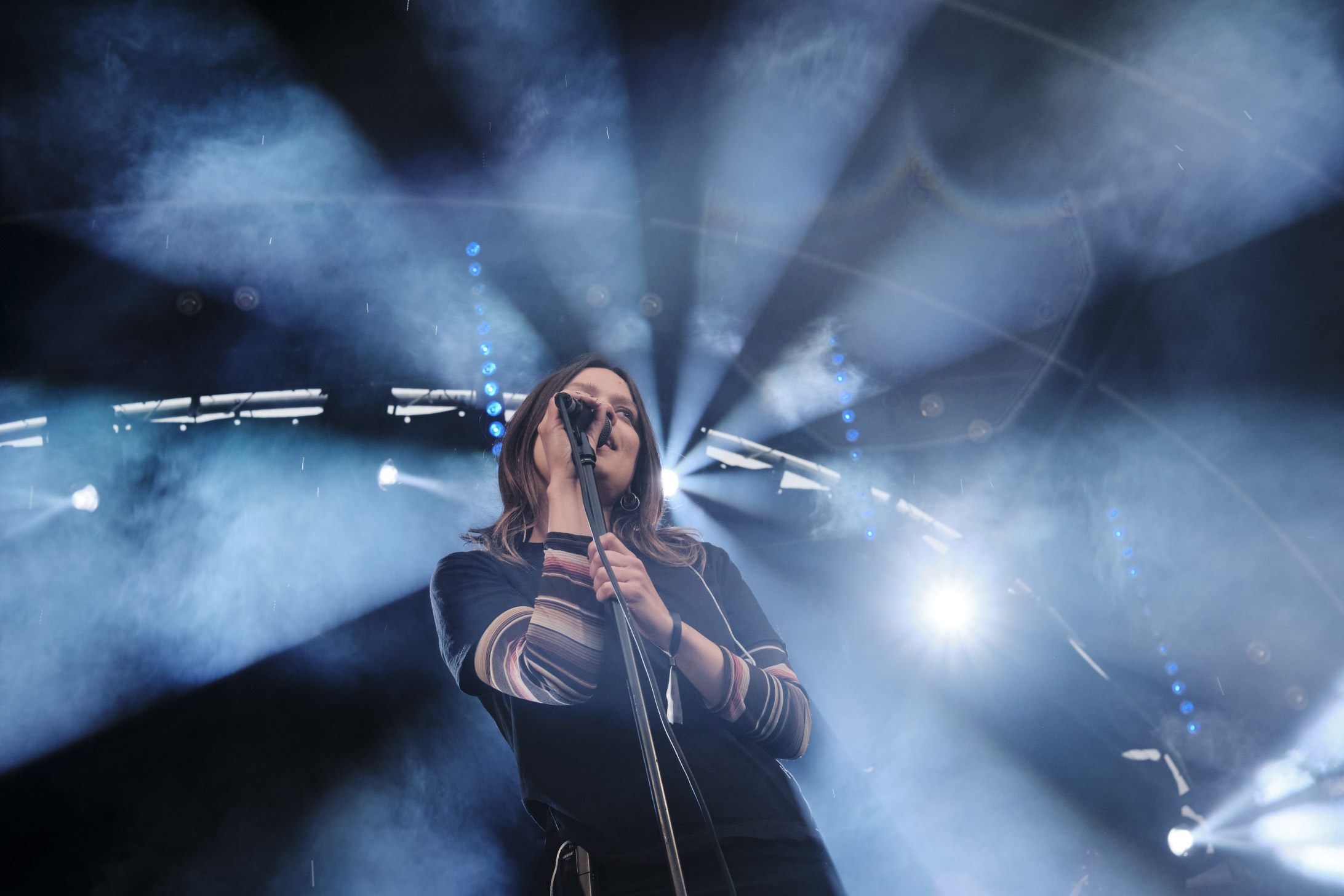 Female singer performing on stage with dramatic blue and white smoke and light beams behind her.