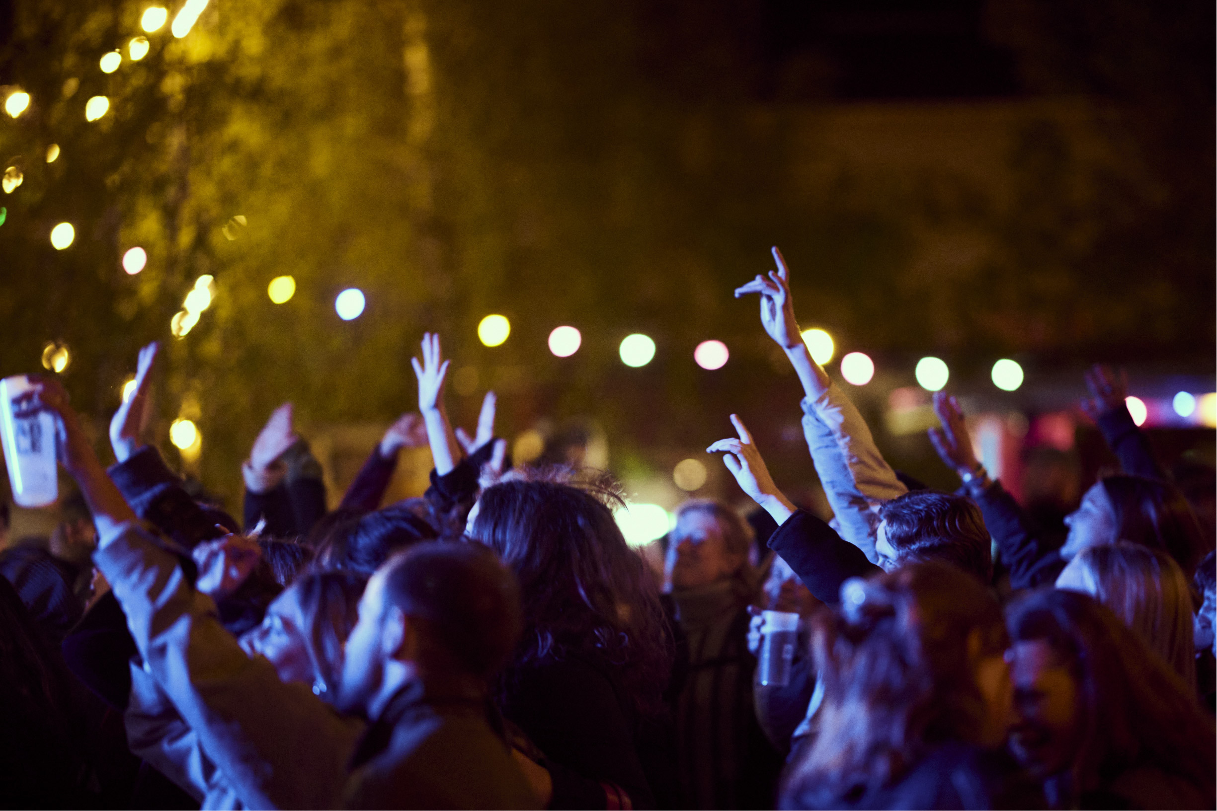 Crowd of people at night raising hands amid colorful string lights in background.