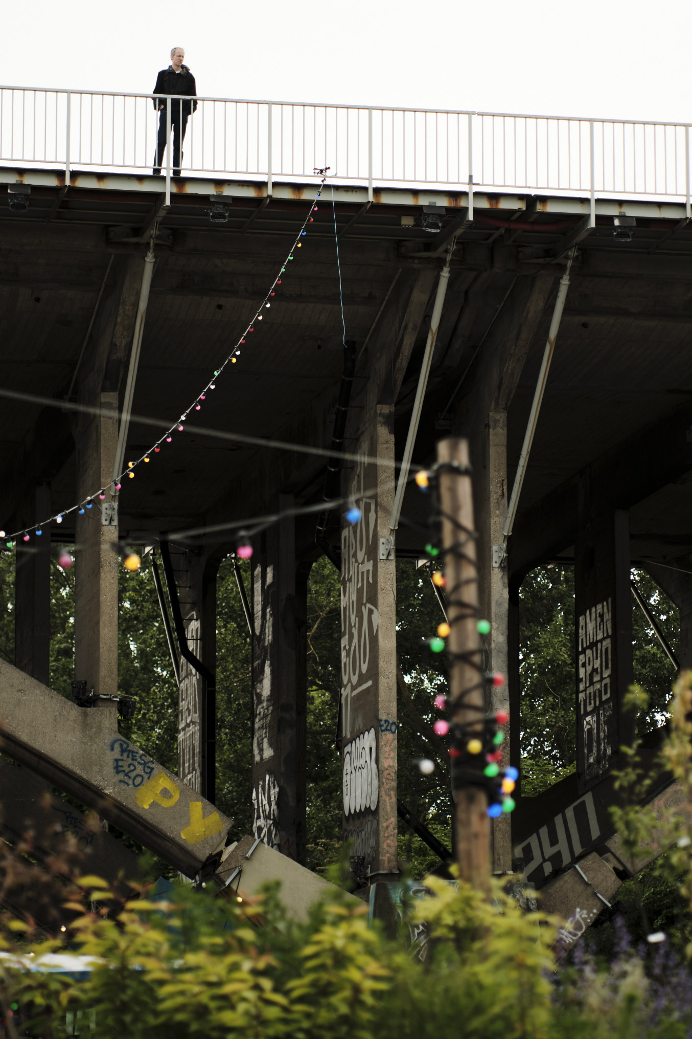 Man standing on a bridge railing above graffiti-covered concrete pillars with string lights below.