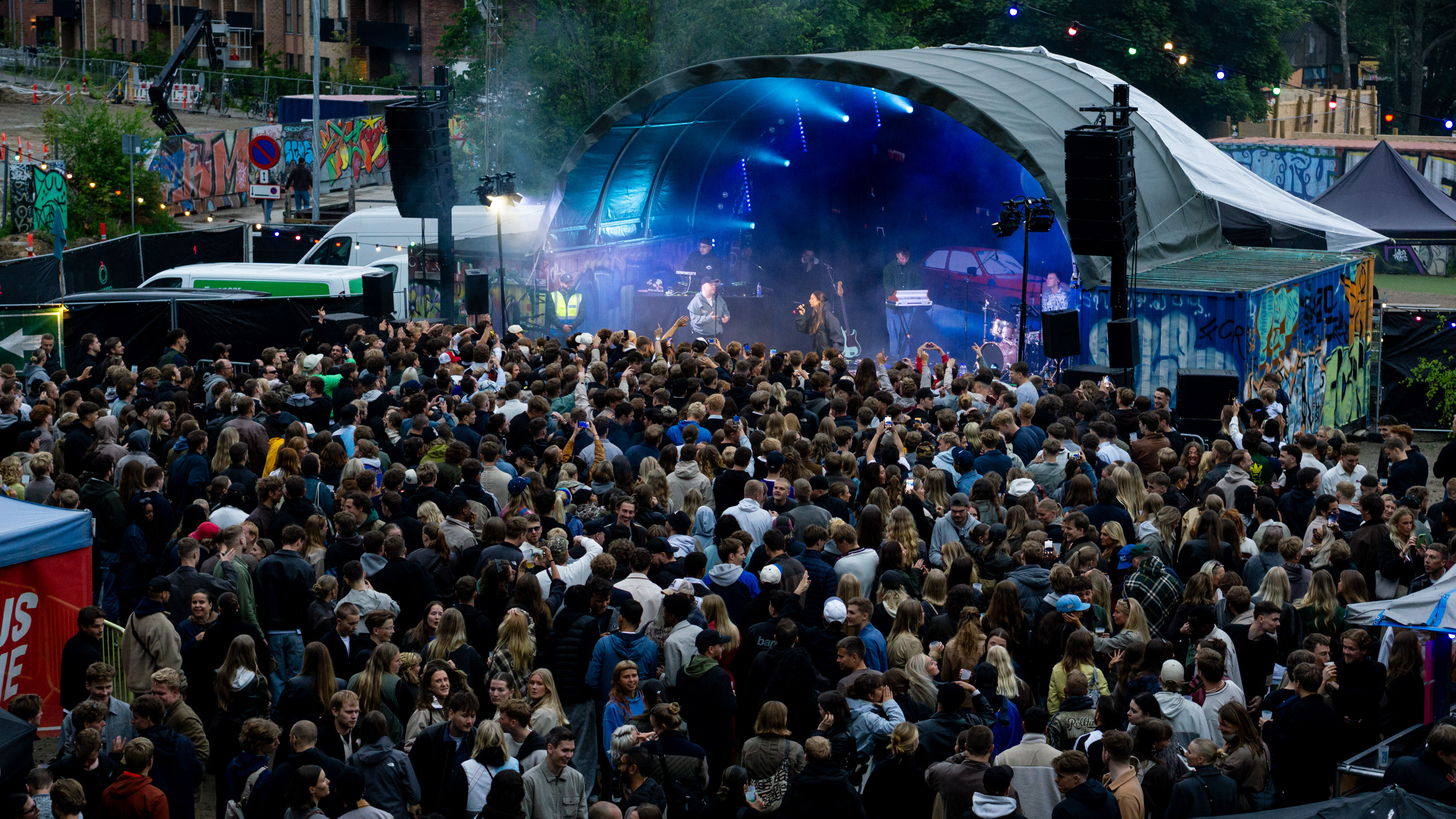 Large crowd watching live musicians performing on outdoor stage with colorful graffiti background.