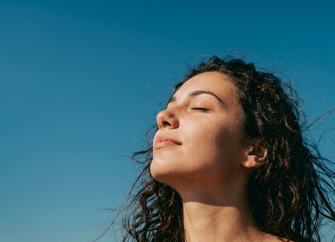 Woman with curly hair facing the sun, eyes closed, enjoying the clear blue sky.