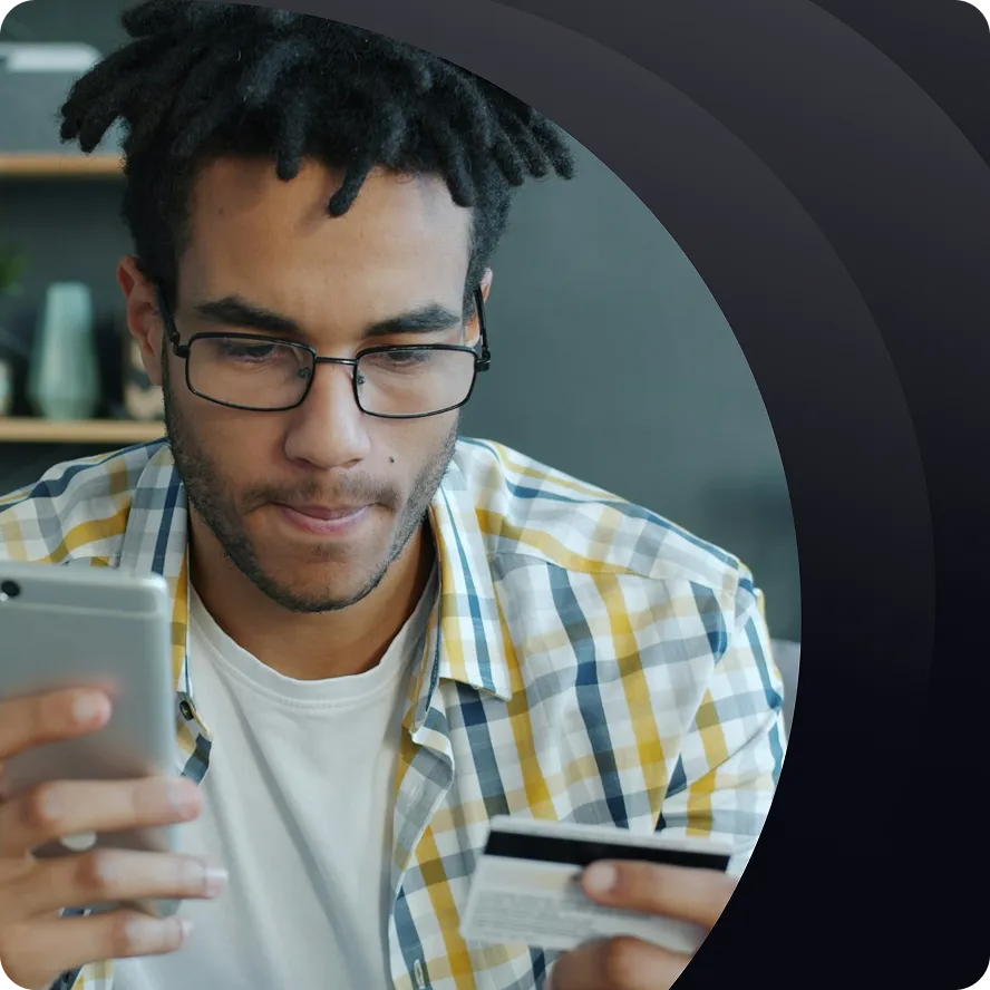 Homme portant des lunettes, tenant une carte bancaire d'une main, un smartphone de l'autre, concentré.