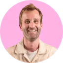Homme souriant avec une chemise beige sur fond rose.